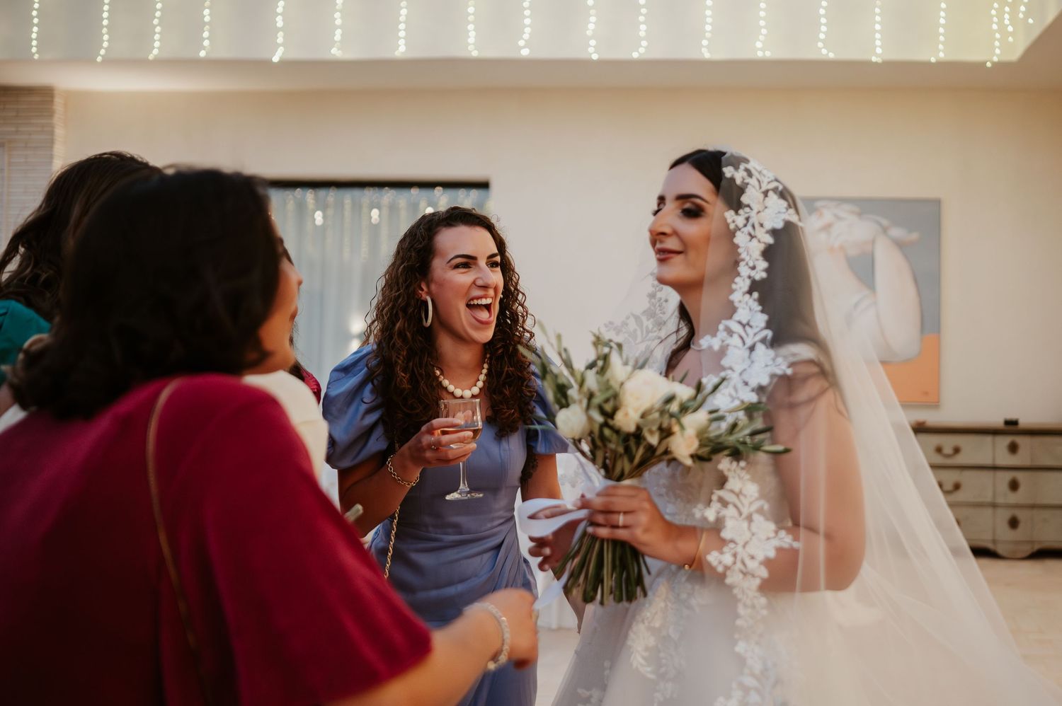 Bride in white gown laughing with guests while holding a bouquet at wedding reception.