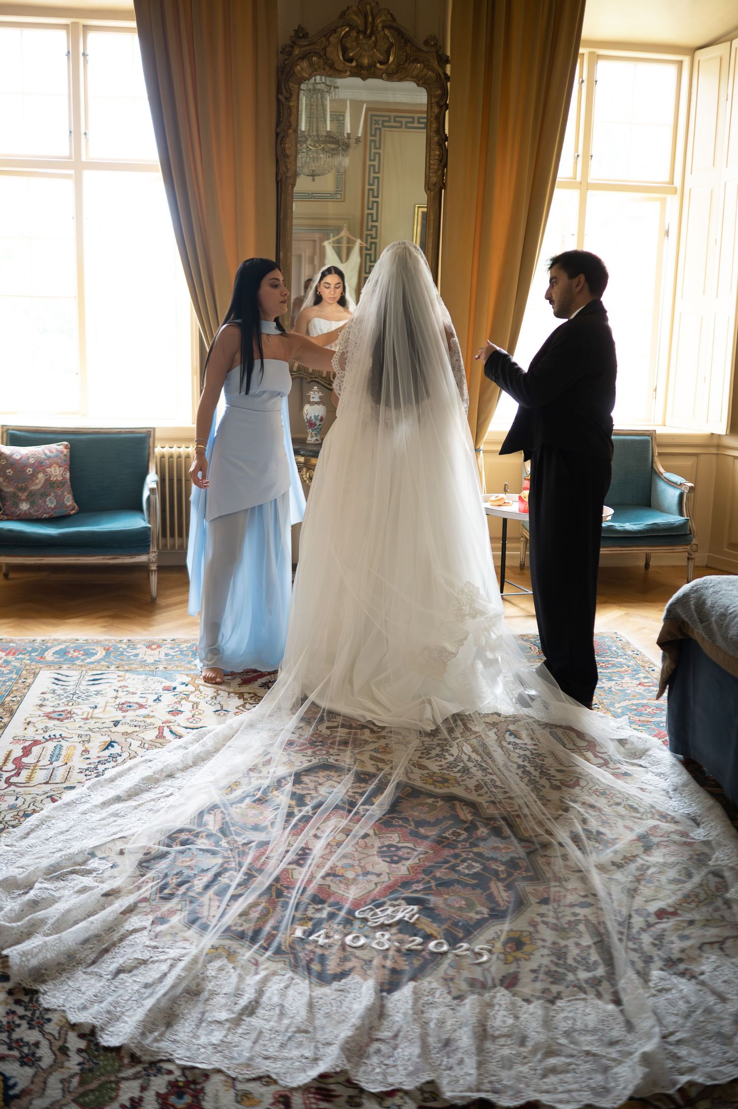 Bride in cathedral-length embroidered veil being attended by bridesmaid and groomsman in ornate room.