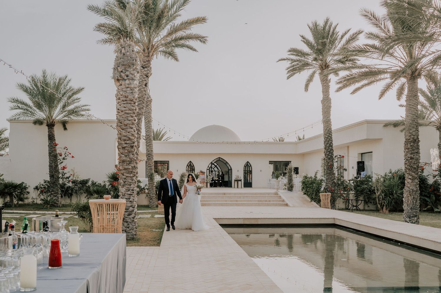 Bride and groom walking hand in hand along a reflecting pool at a stunning palm-tree-lined wedding venue.