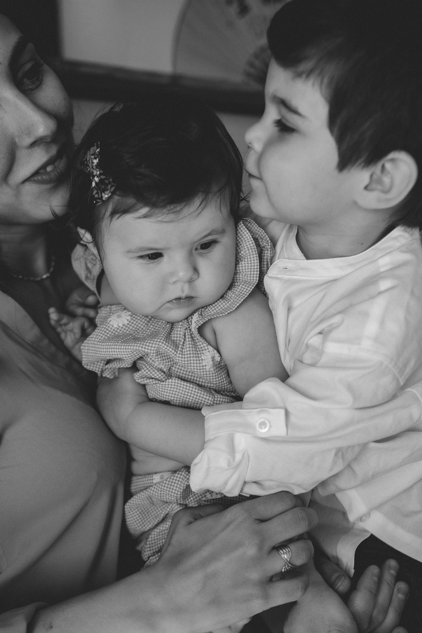 Black and white photo of a mother with two children, older sibling kissing baby on the cheek tenderly.