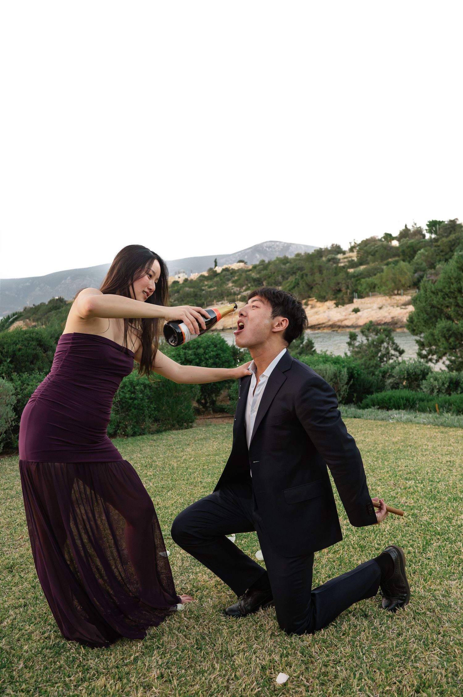 Playful wedding guests in garden, woman pouring champagne into kneeling mans mouth at outdoor reception.