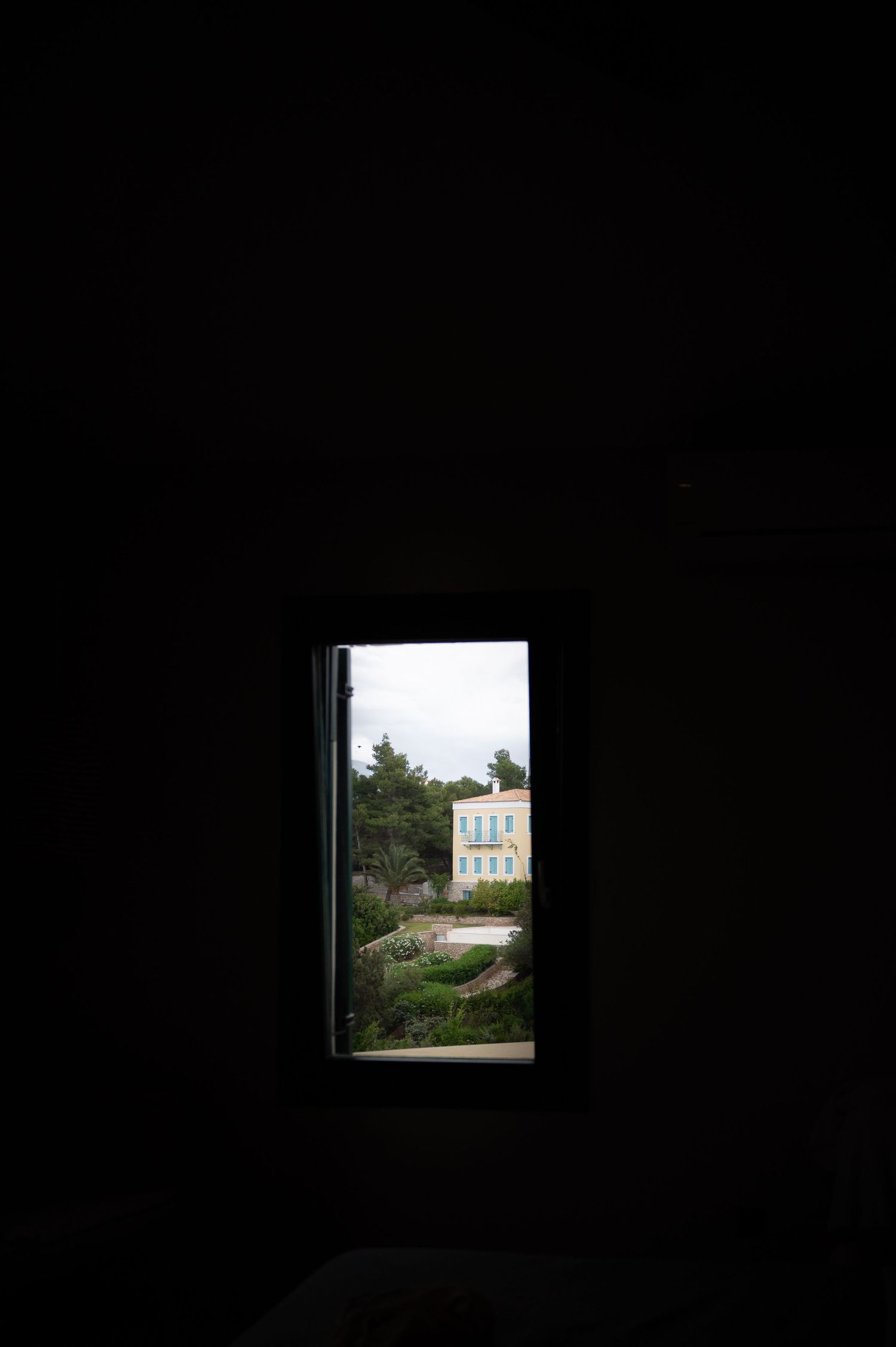 Dark room with a small window revealing a yellow building surrounded by trees and gardens outside.