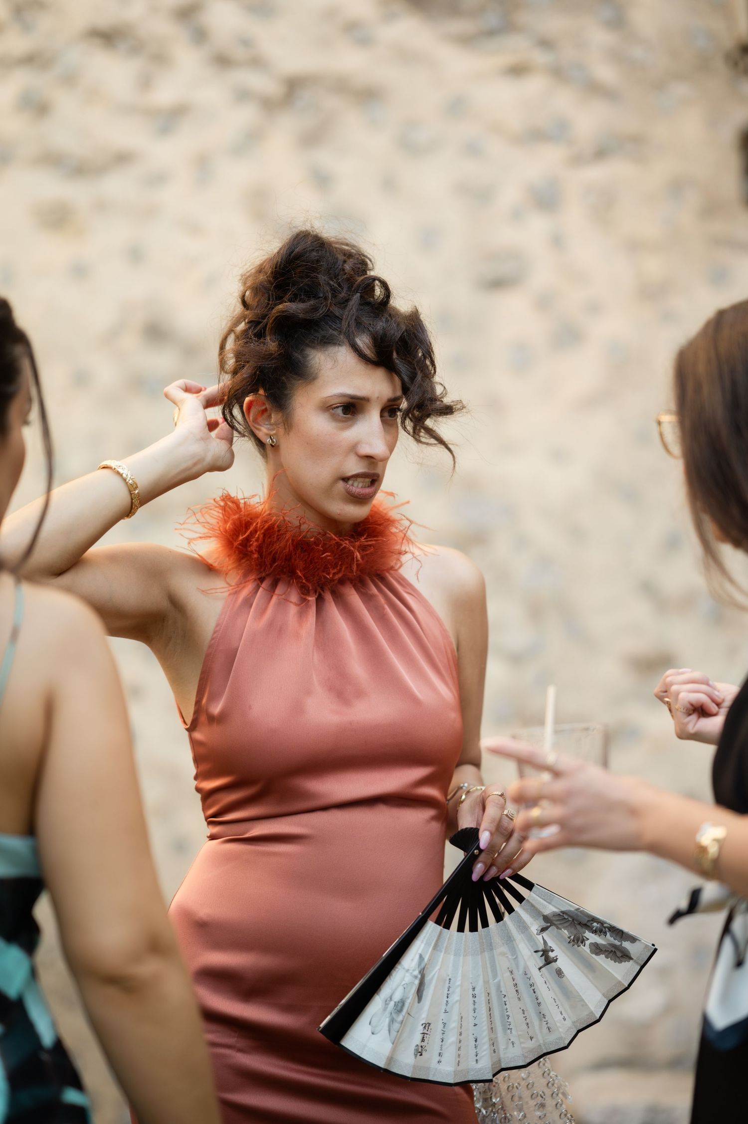 Woman in dusty rose halter gown with feather neckline holds a decorative fan at an outdoor wedding reception.