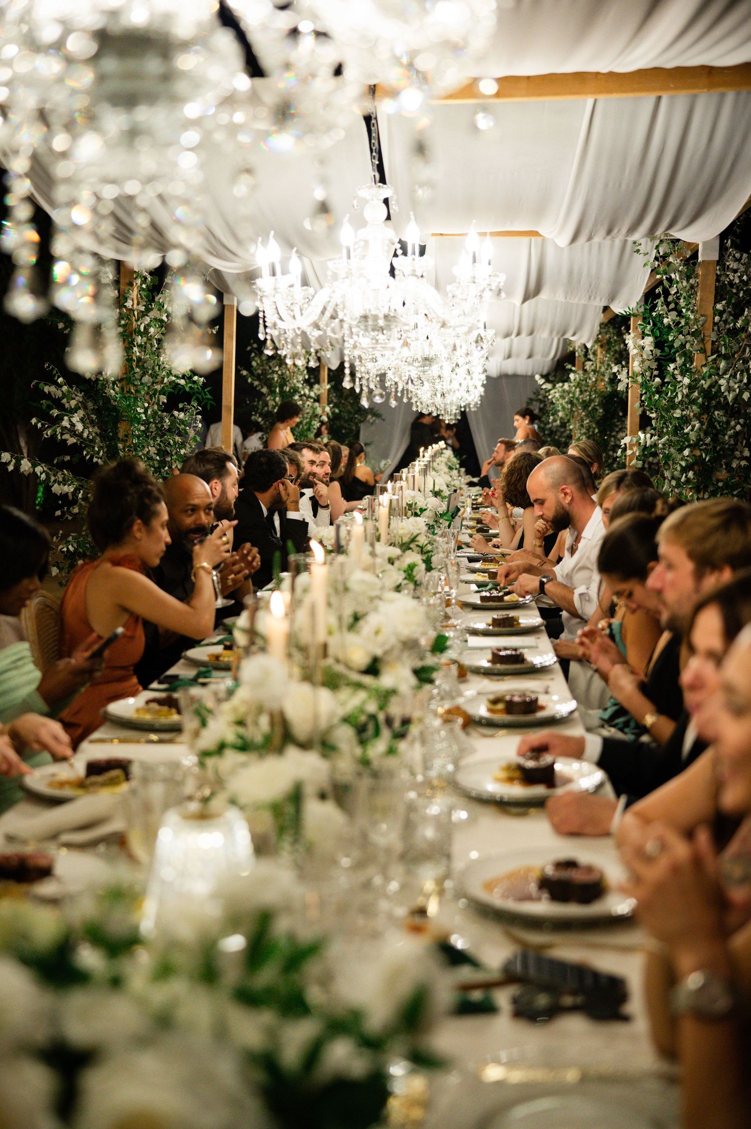 Luxurious wedding reception with long banquet table, crystal chandeliers, candlelight, and lush greenery decor.