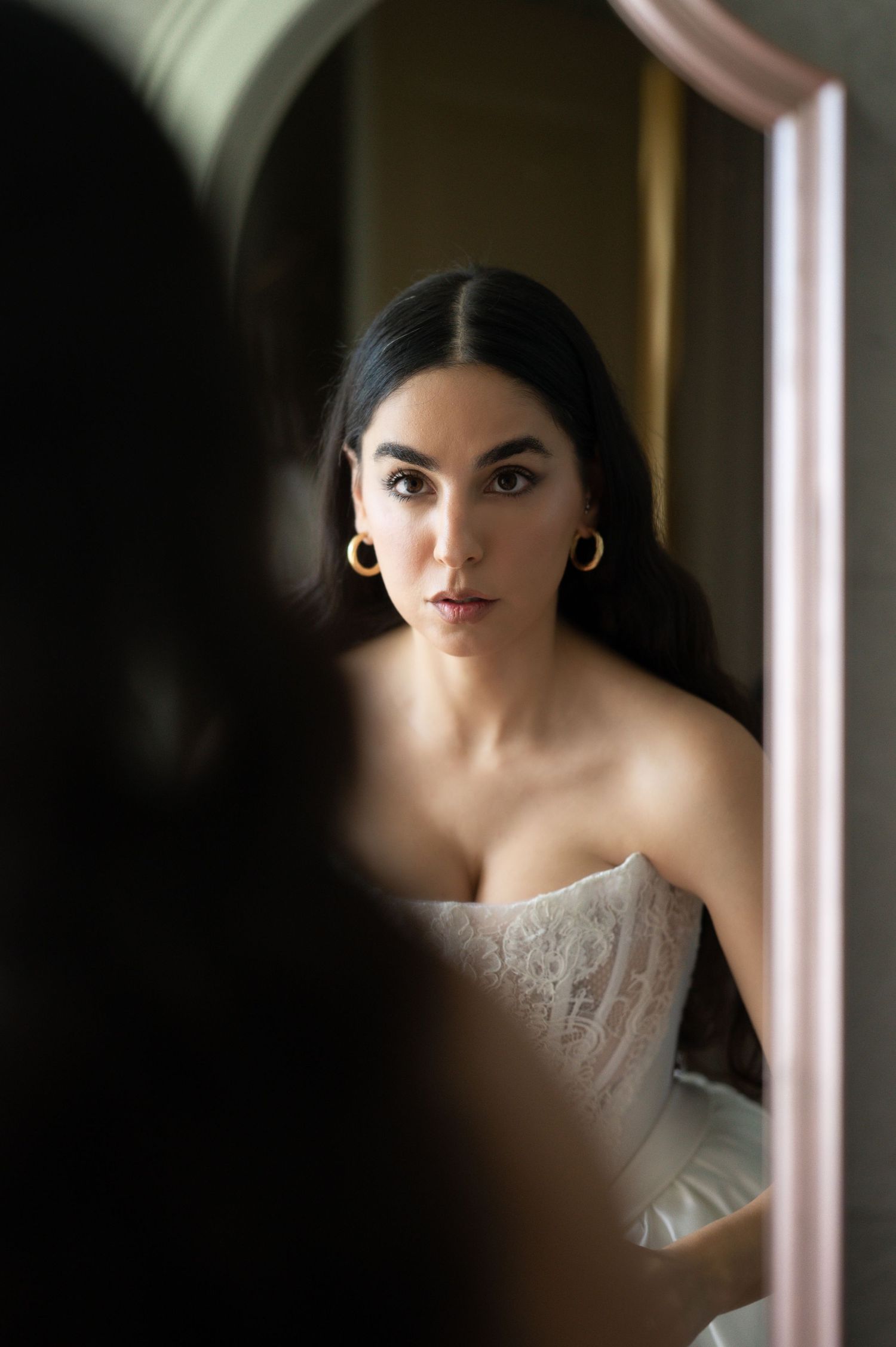 Dark-haired bride in white lace corset gazing intensely at her reflection in a pink-framed mirror.