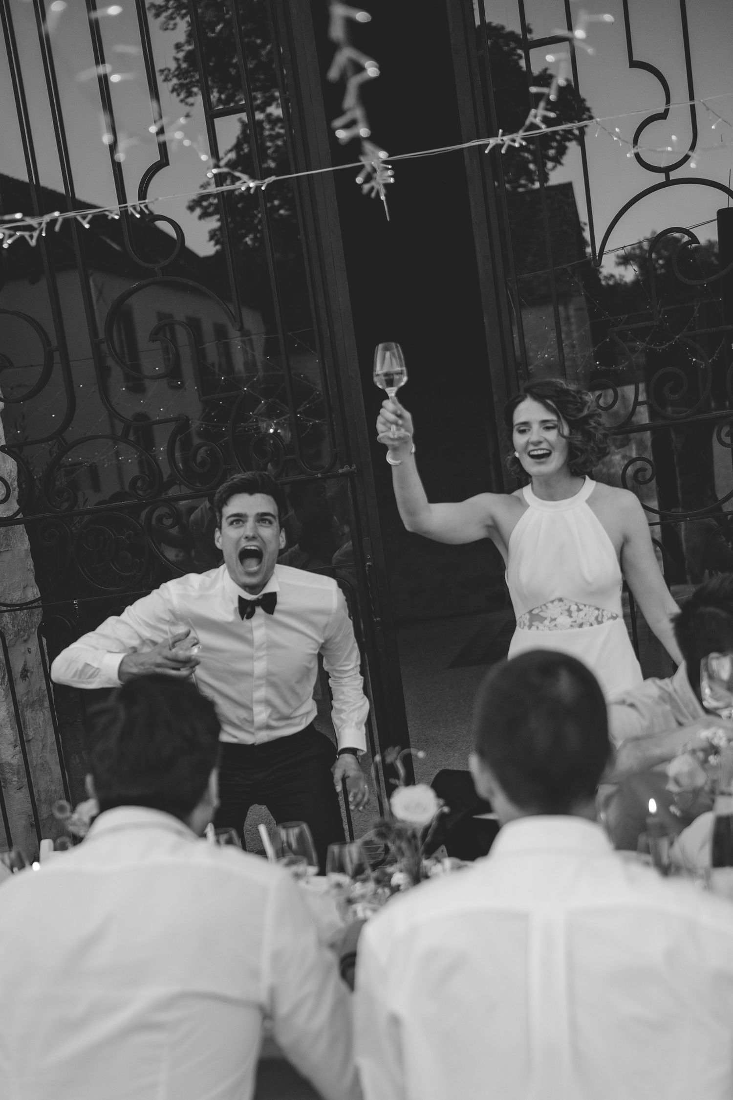Joyful bride raises champagne glass as groom cheers during lively wedding reception.