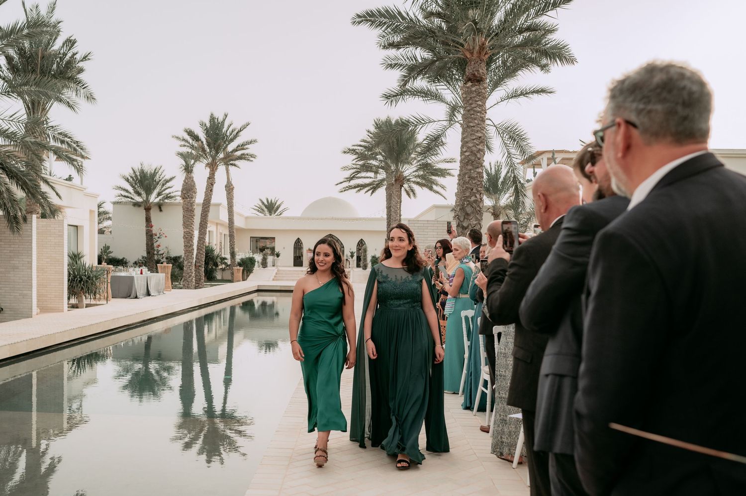 Bridesmaids in emerald green gowns walking along a reflective pool lined with palm trees at an outdoor wedding.