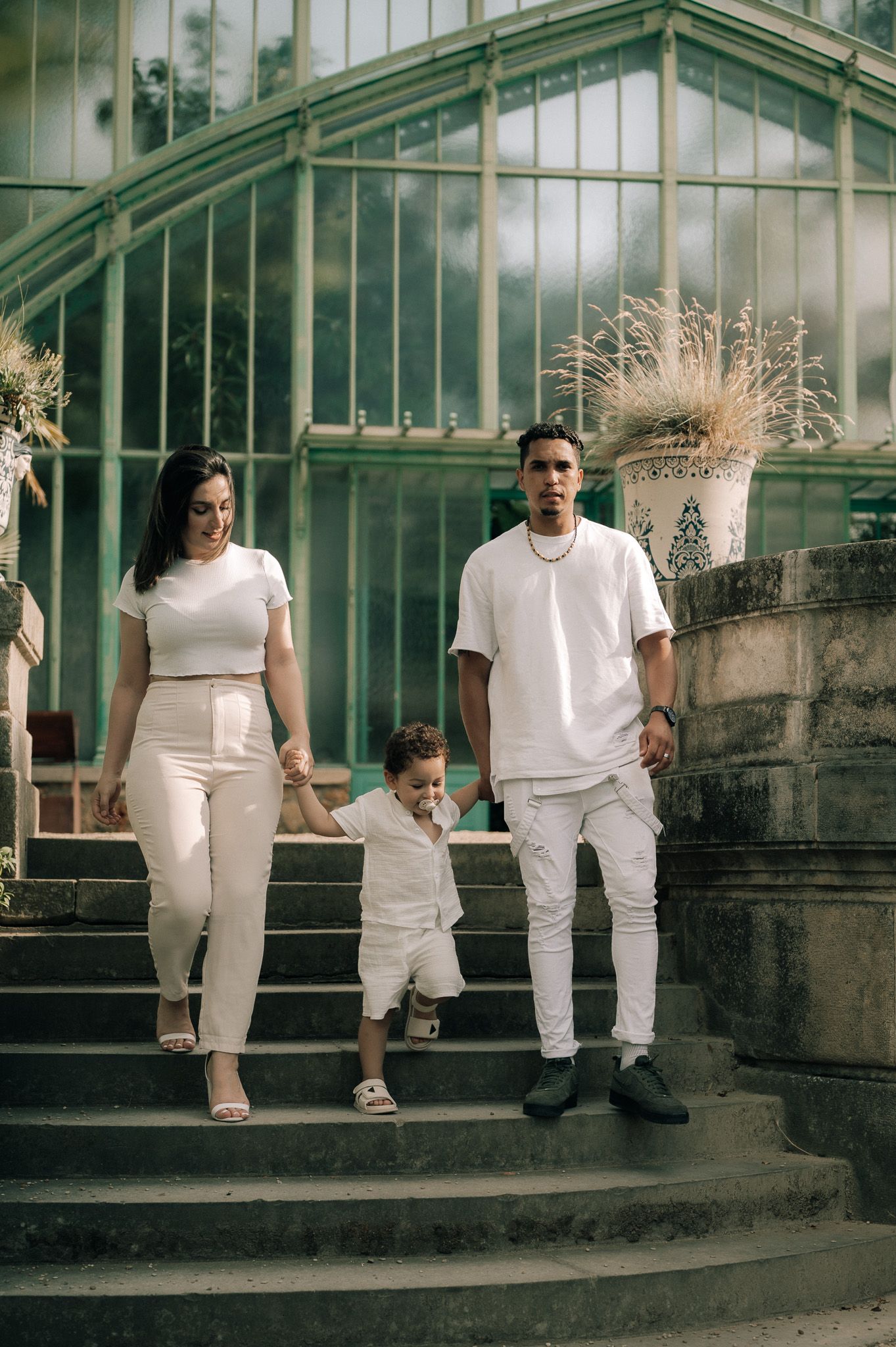 Family of three in matching white outfits descending stone steps in front of a vintage glass greenhouse.