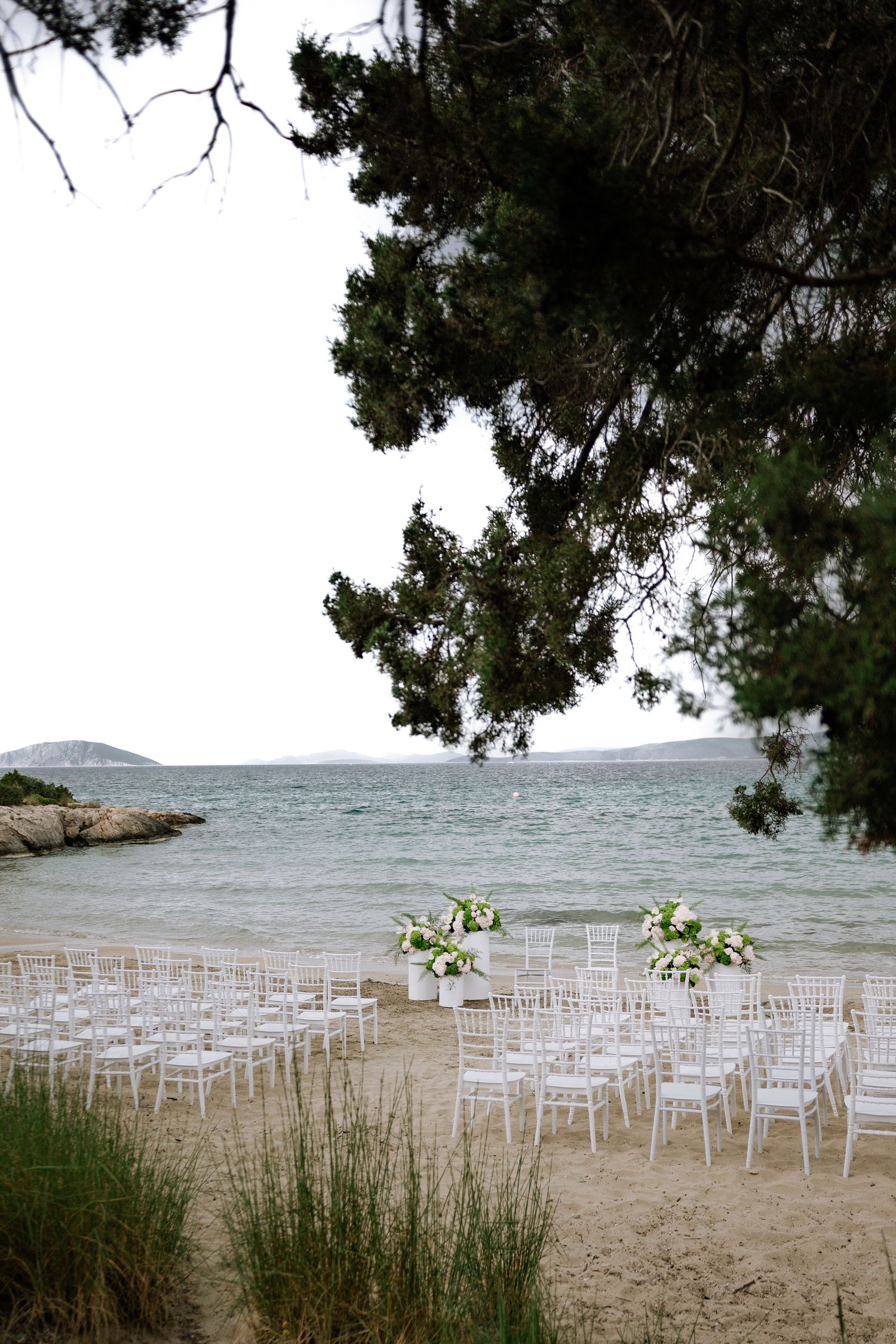 White chairs arranged on a sandy beach for an outdoor wedding ceremony with floral arrangements and ocean views.