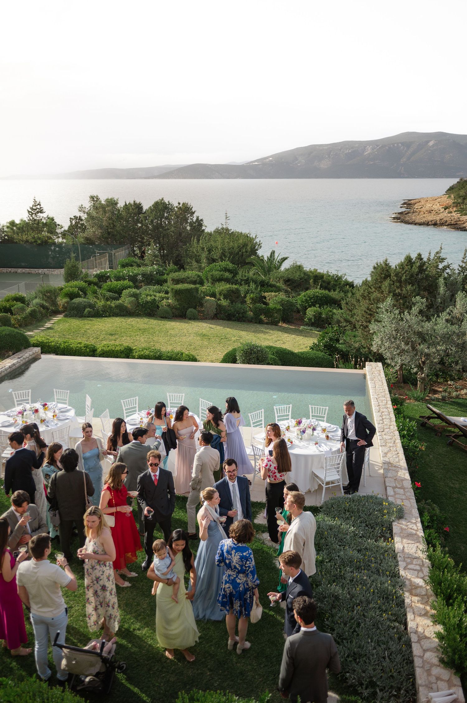 Wedding guests mingle by infinity pool overlooking scenic coastal waters at sunset.