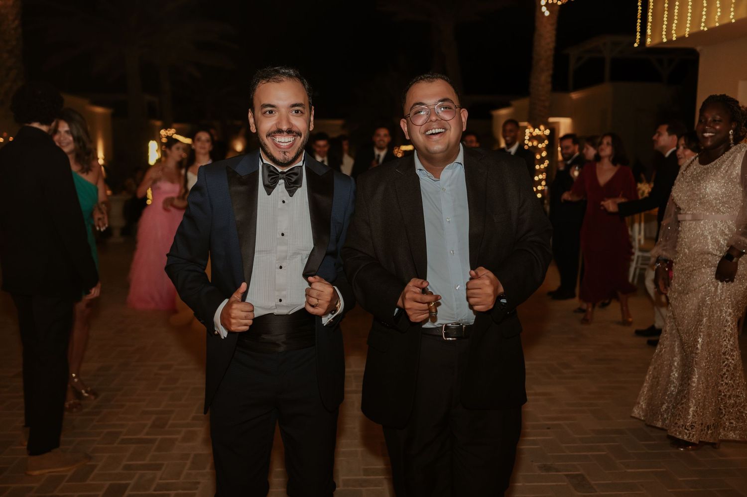 Two men in tuxedos smile and pose together at an outdoor wedding reception at night.