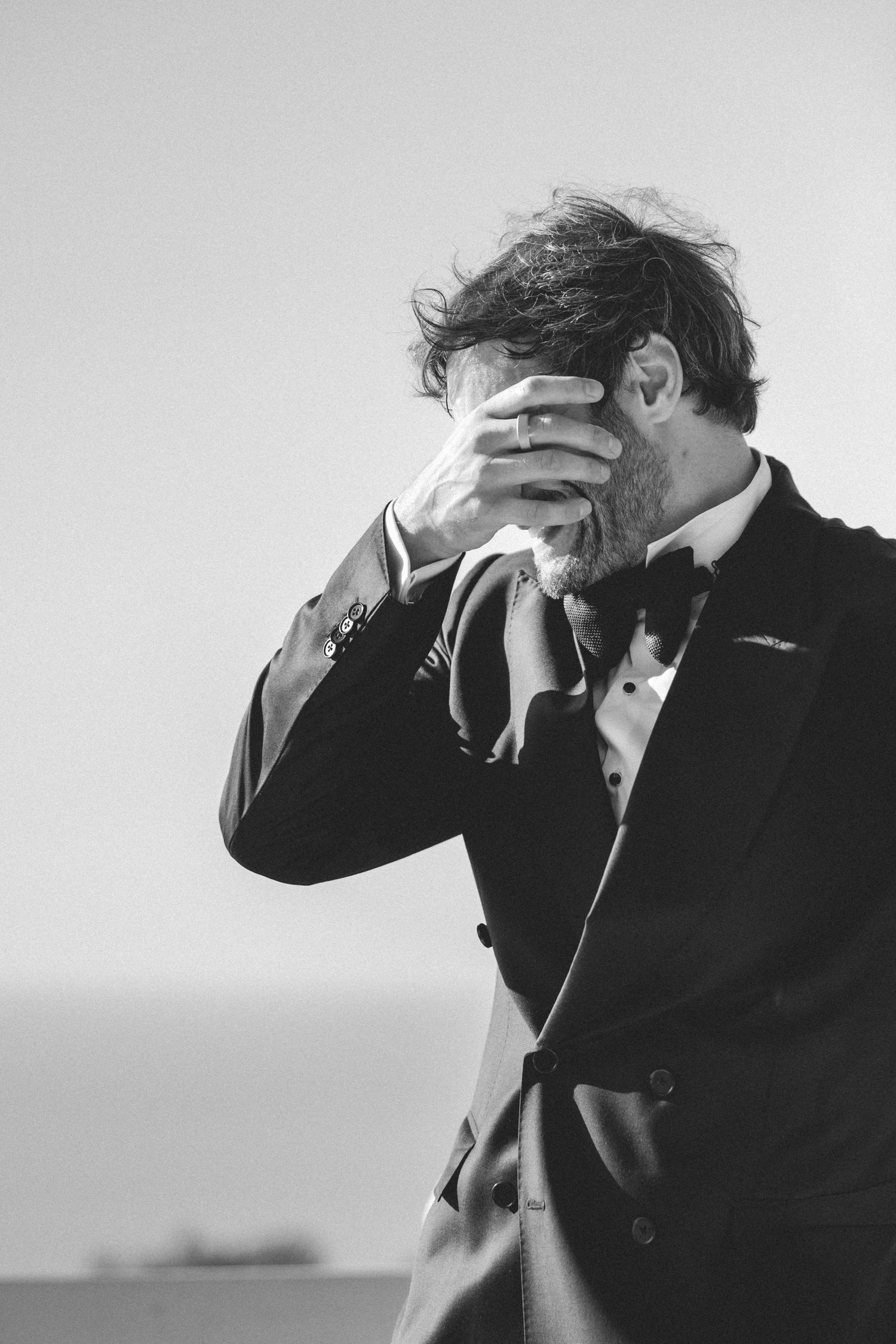 Emotional groom in black tuxedo covers his face with his hand during an outdoor wedding ceremony.