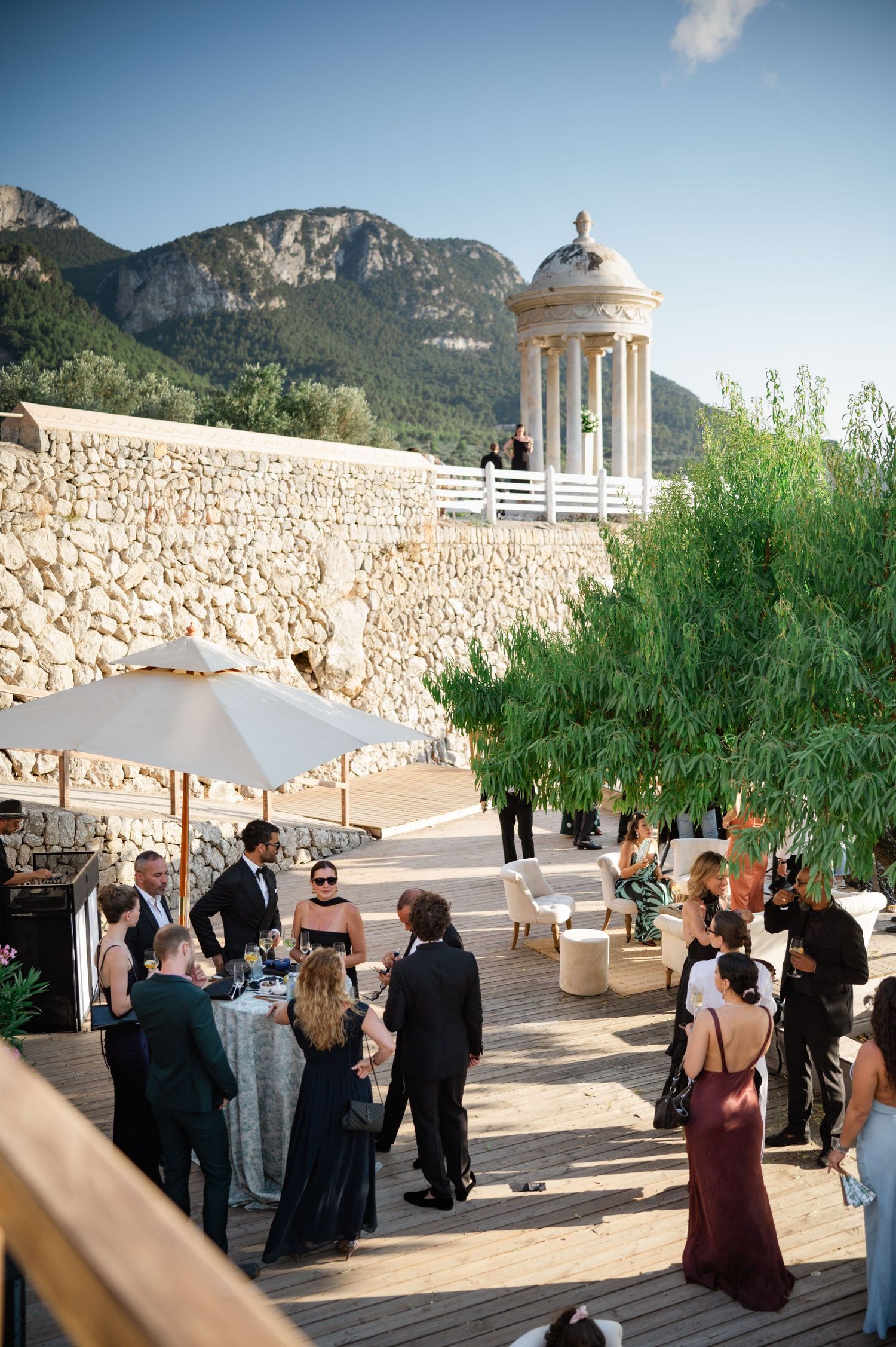 Elegant outdoor wedding cocktail hour on a wooden terrace with stone walls and mountain backdrop in Mallorca.