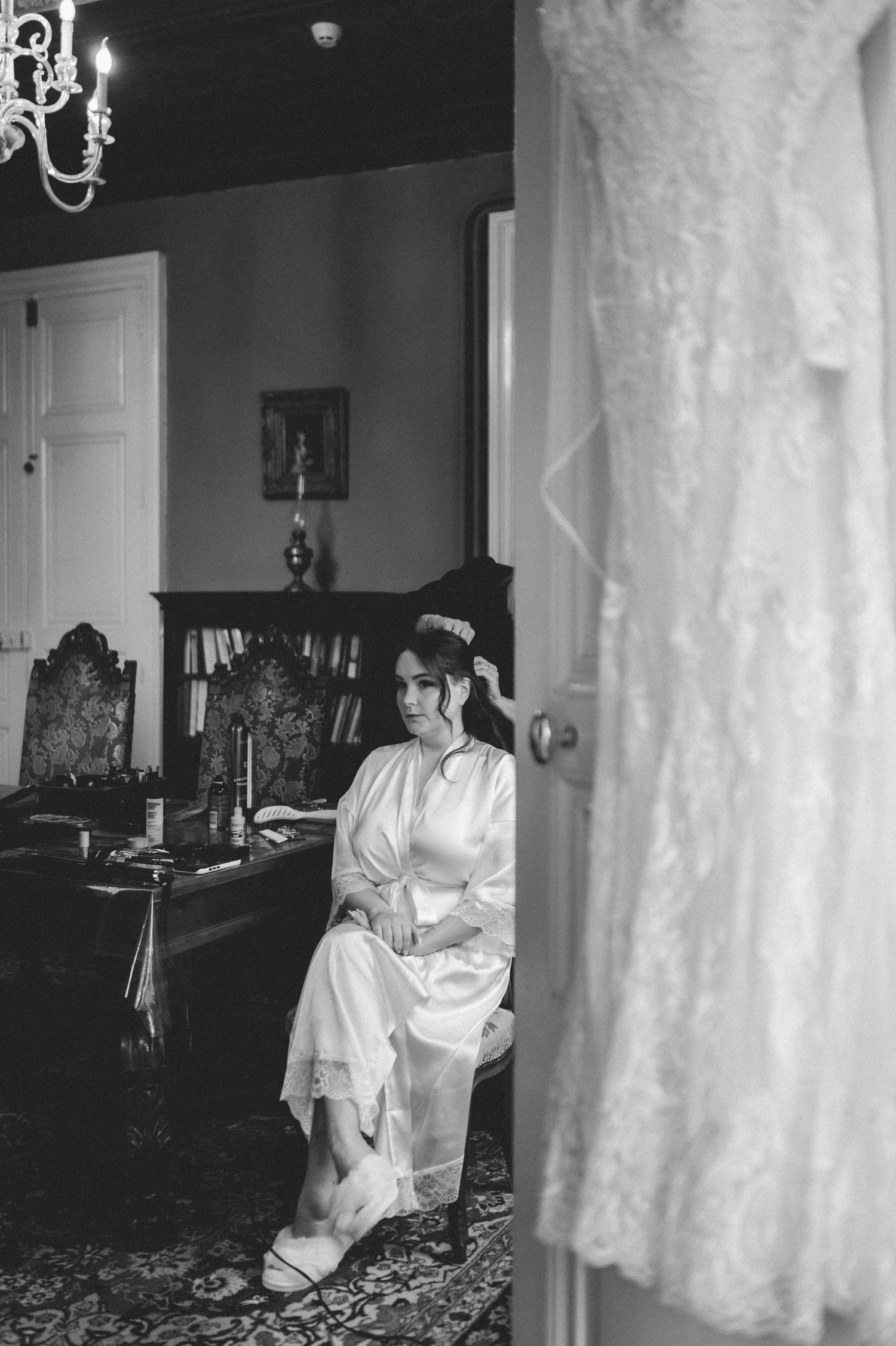 Bride in white robe sits pensively beside her lace wedding gown hanging on a door in an elegant room.
