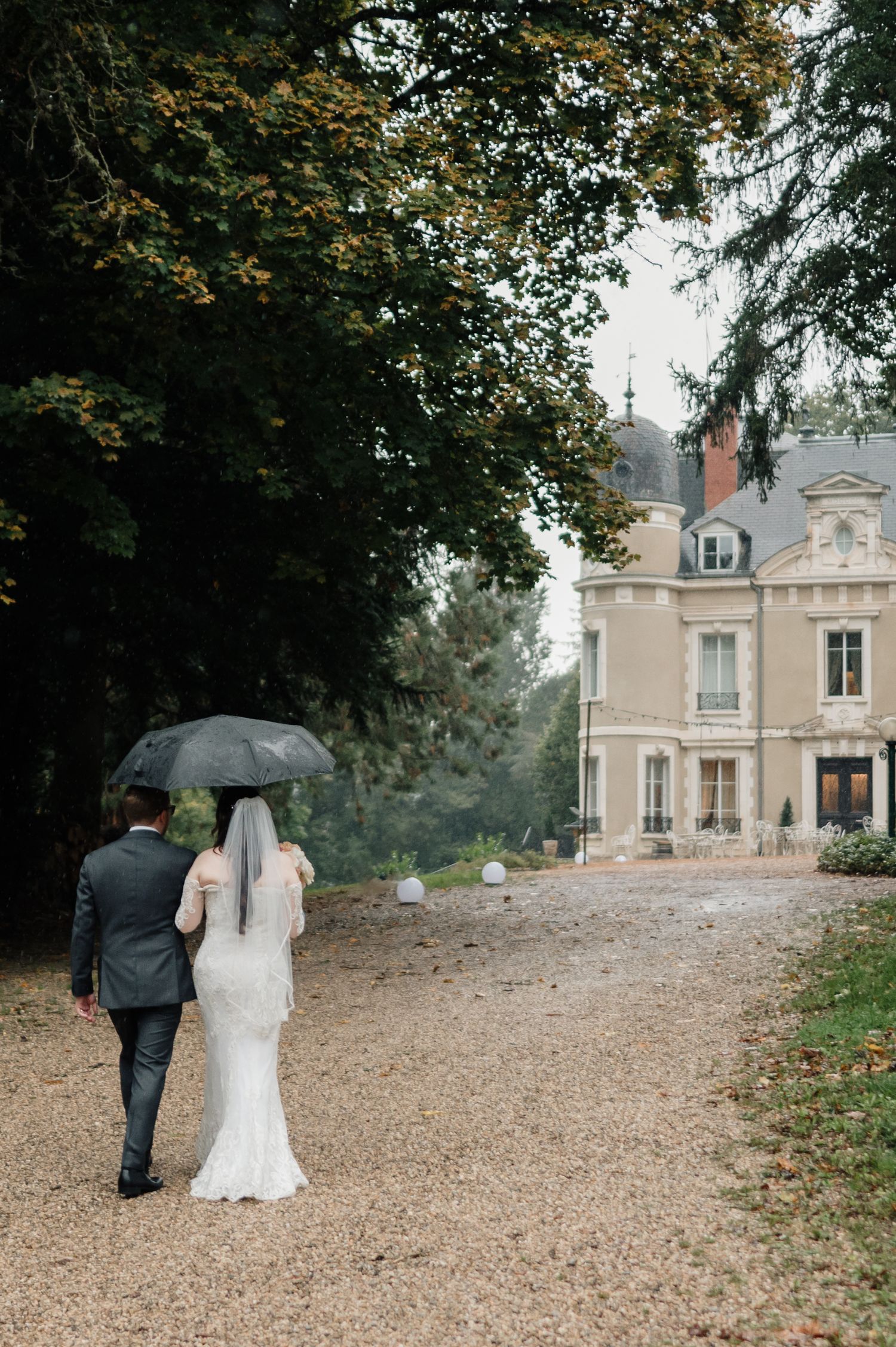 Bride and groom walk under umbrella along gravel path toward elegant French chateau in rain.