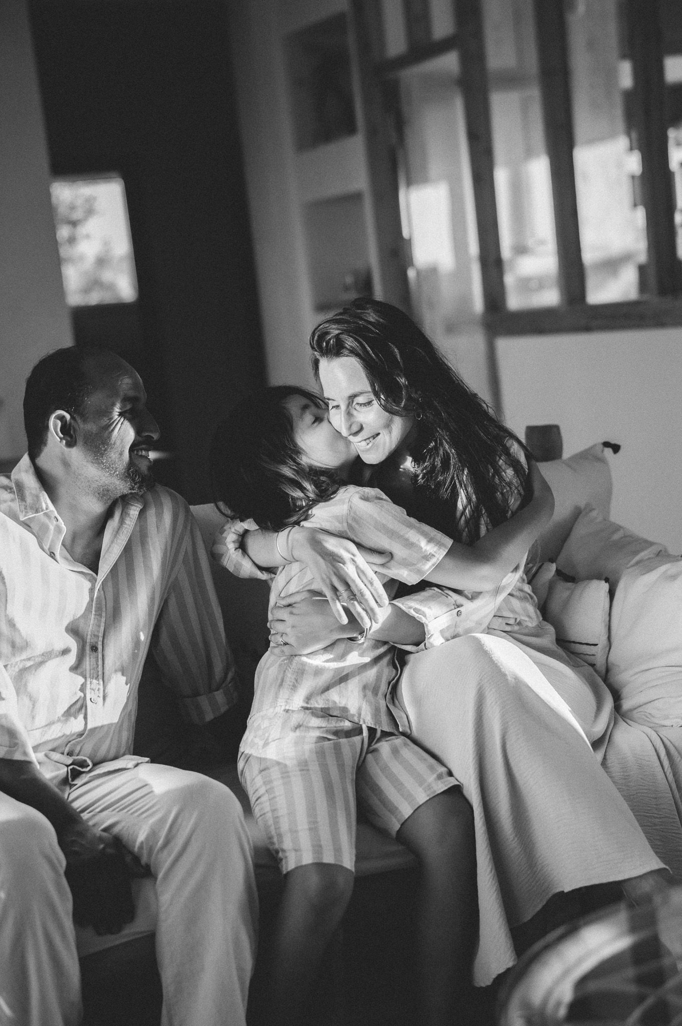 Black and white photo of young boy kissing mother on cheek while father watches on sofa.