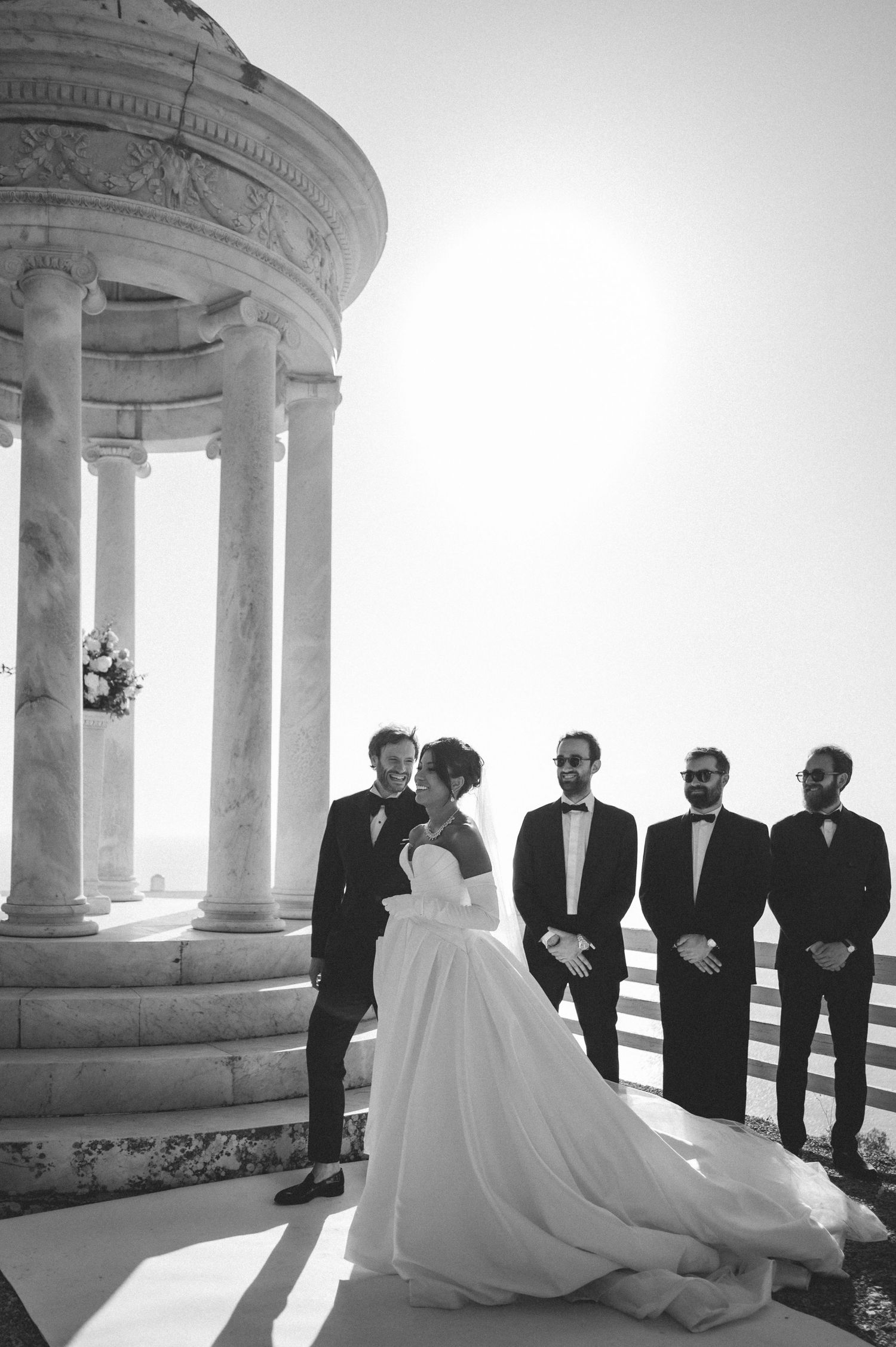Black and white photo of newlyweds sharing a moment by a classical marble rotunda with groomsmen nearby.