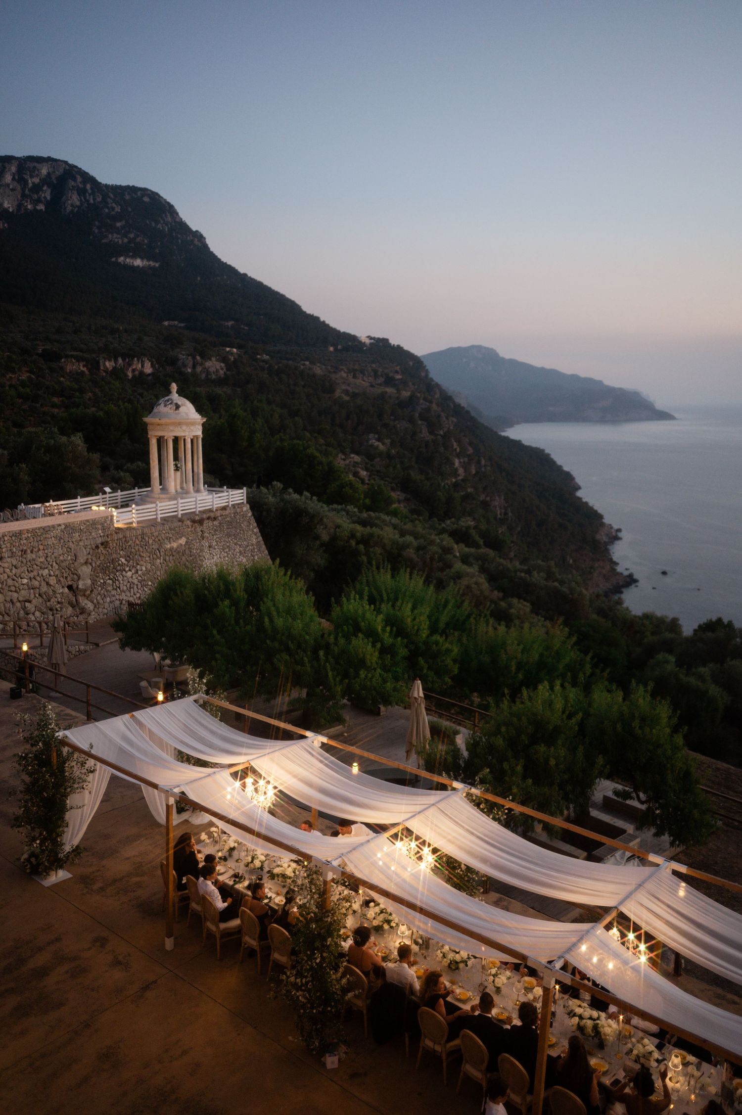 Aerial view of stunning cliffside wedding reception with draped canopy overlooking Mediterranean sea.