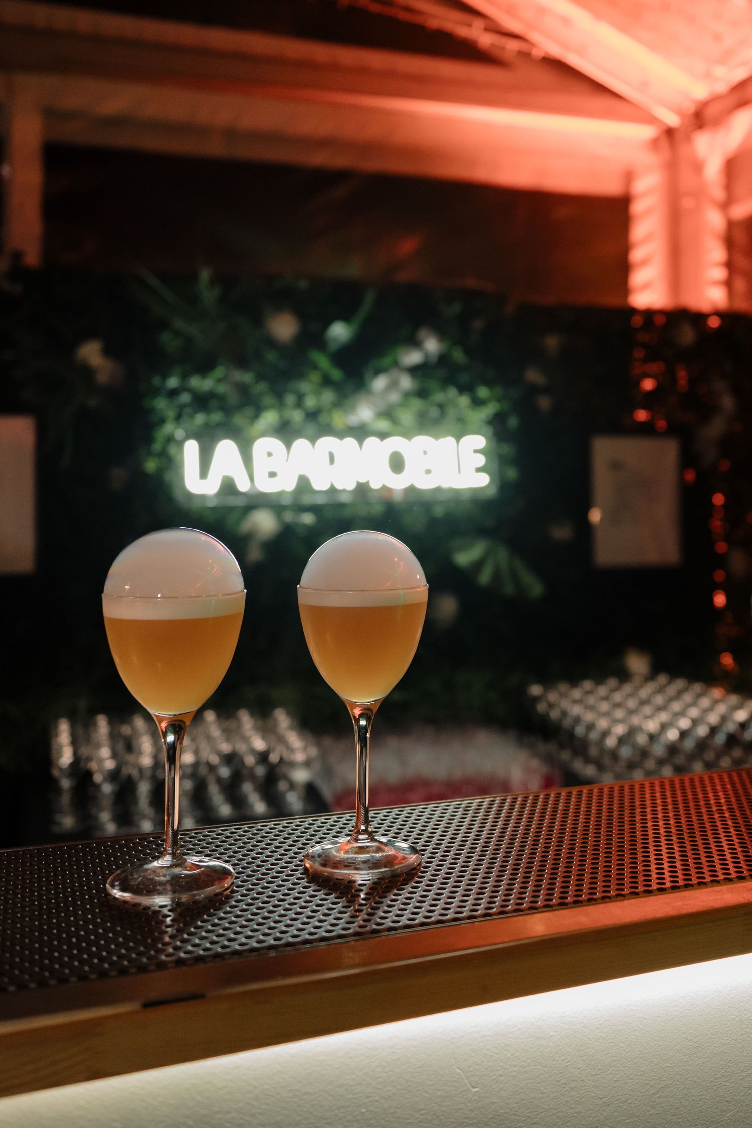 Two elegant cocktails on bar counter with La Barmobile neon sign glowing in background.