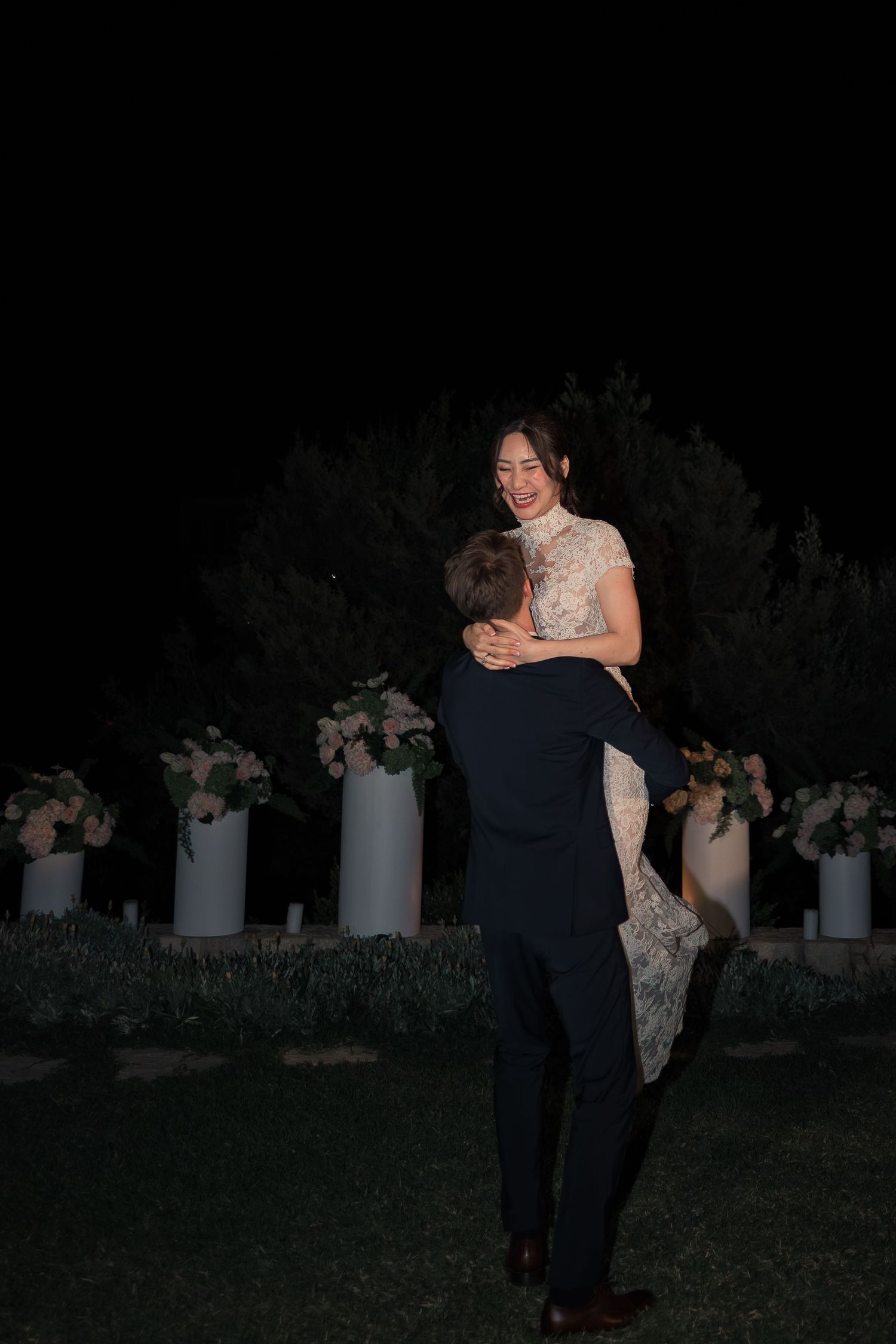Groom lifting smiling bride joyfully during outdoor wedding reception at night.
