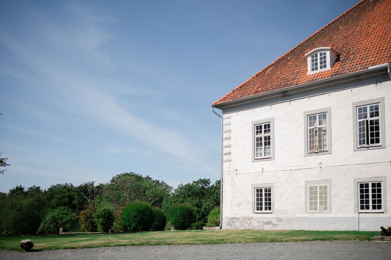 Elegant white manor house with red tile roof surrounded by lush green trees under blue sky.