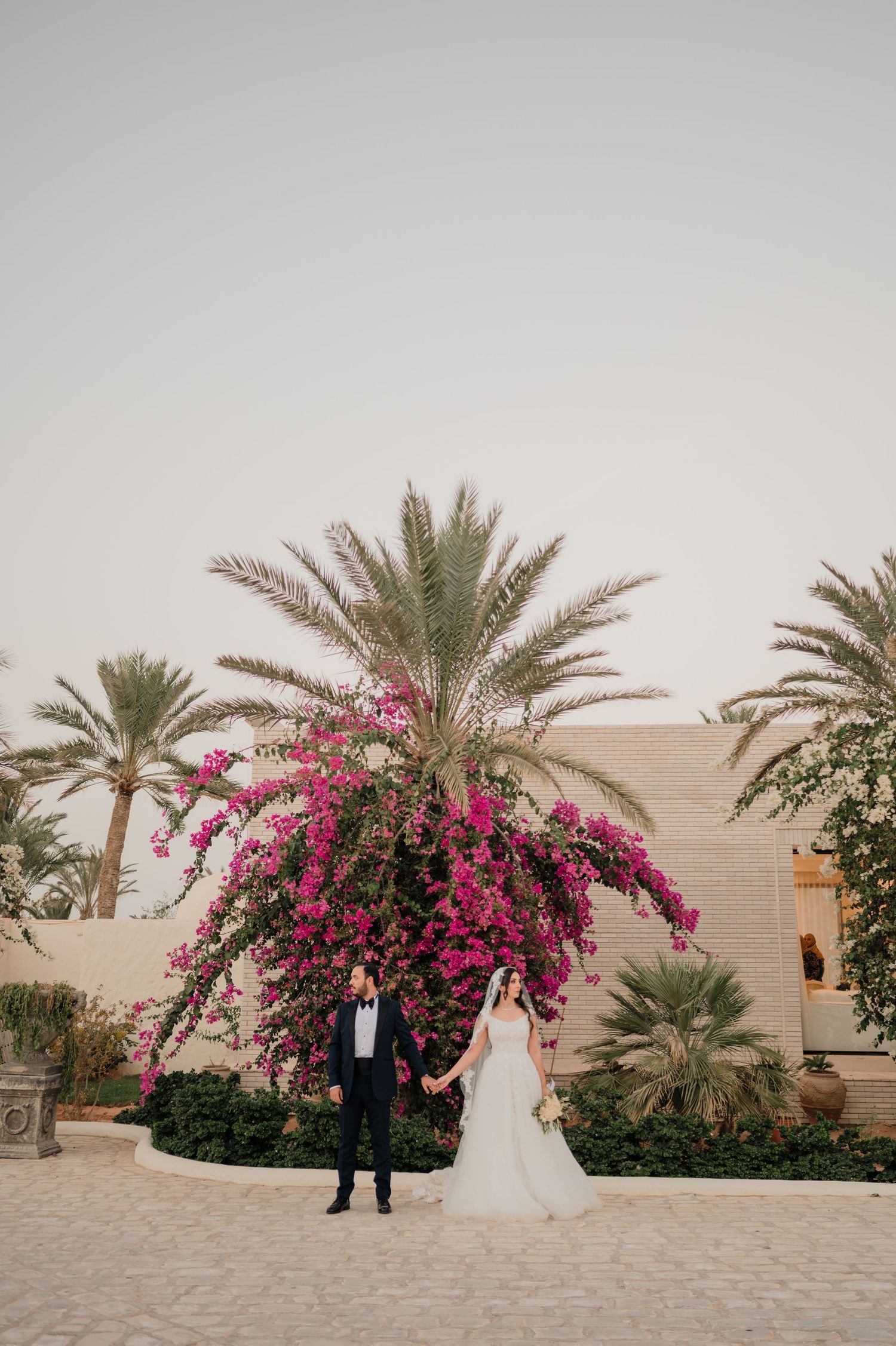Bride and groom holding hands before vibrant pink bougainvillea and tall palm trees at outdoor wedding venue.