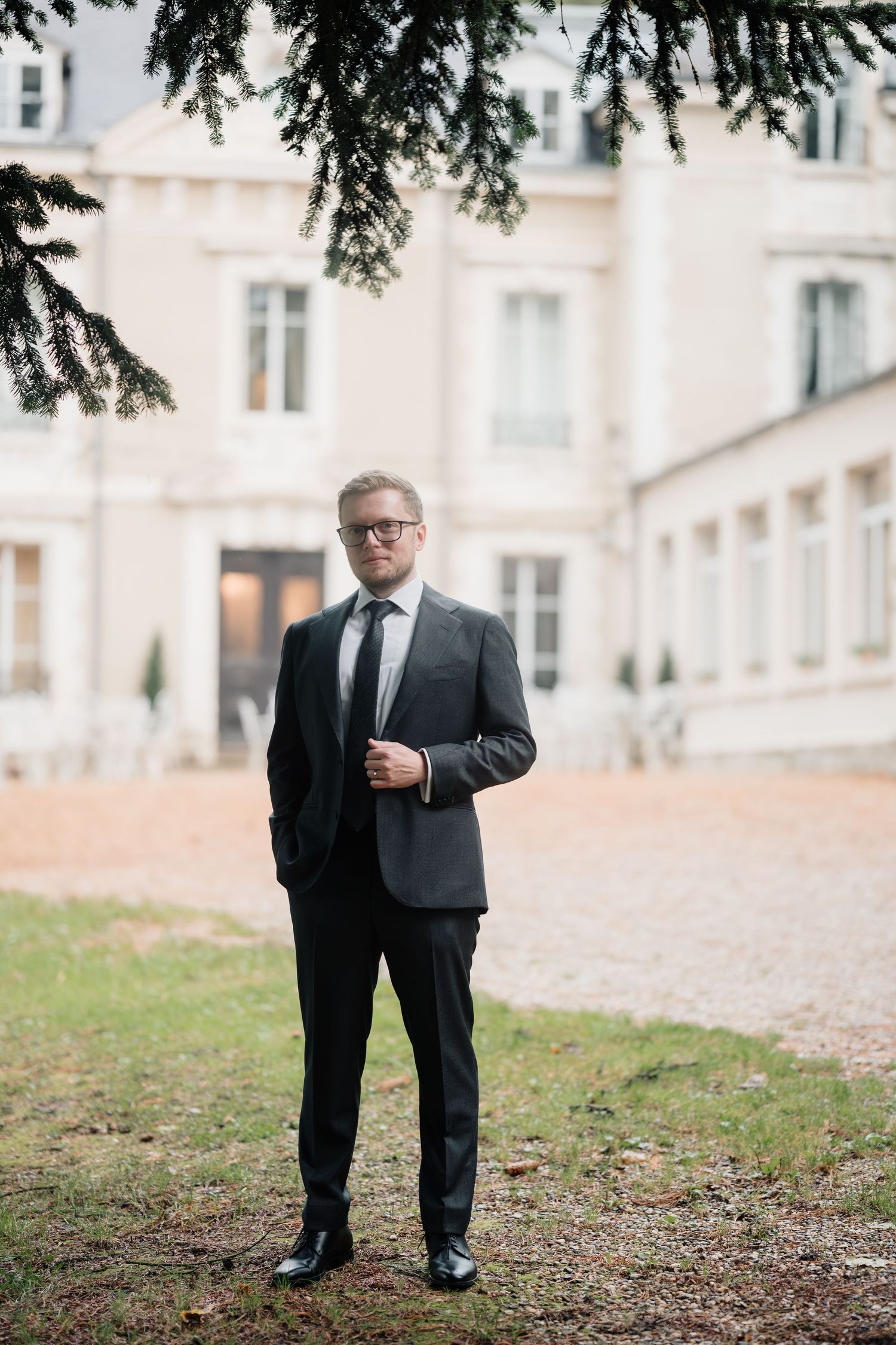 Groom in dark suit and glasses poses confidently outside elegant French chateau wedding venue.