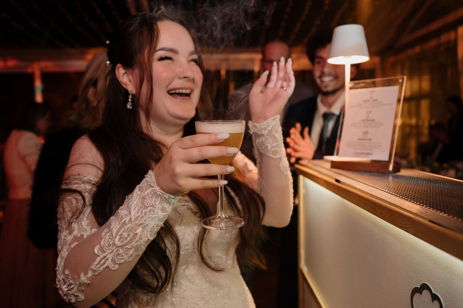 Smiling bride in lace gown holds cocktail at wedding reception bar.