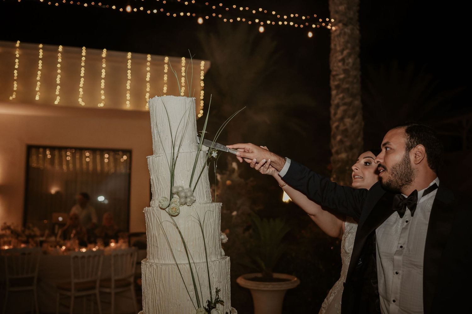 Bride and groom cutting tall four-tier white wedding cake outdoors under string lights at night.