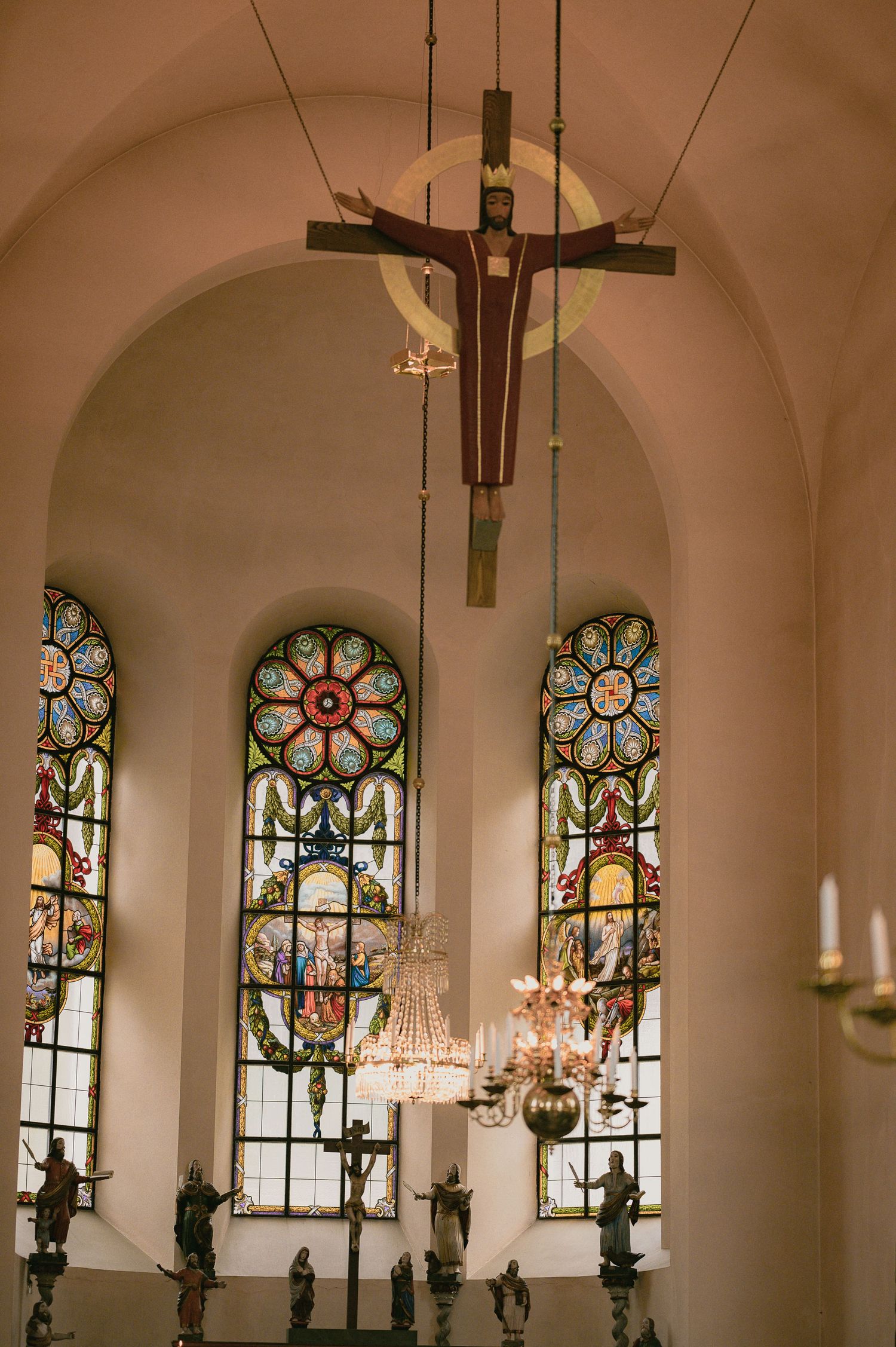 Church interior with crucifix, colorful stained glass windows, chandelier, and religious figurines on altar.