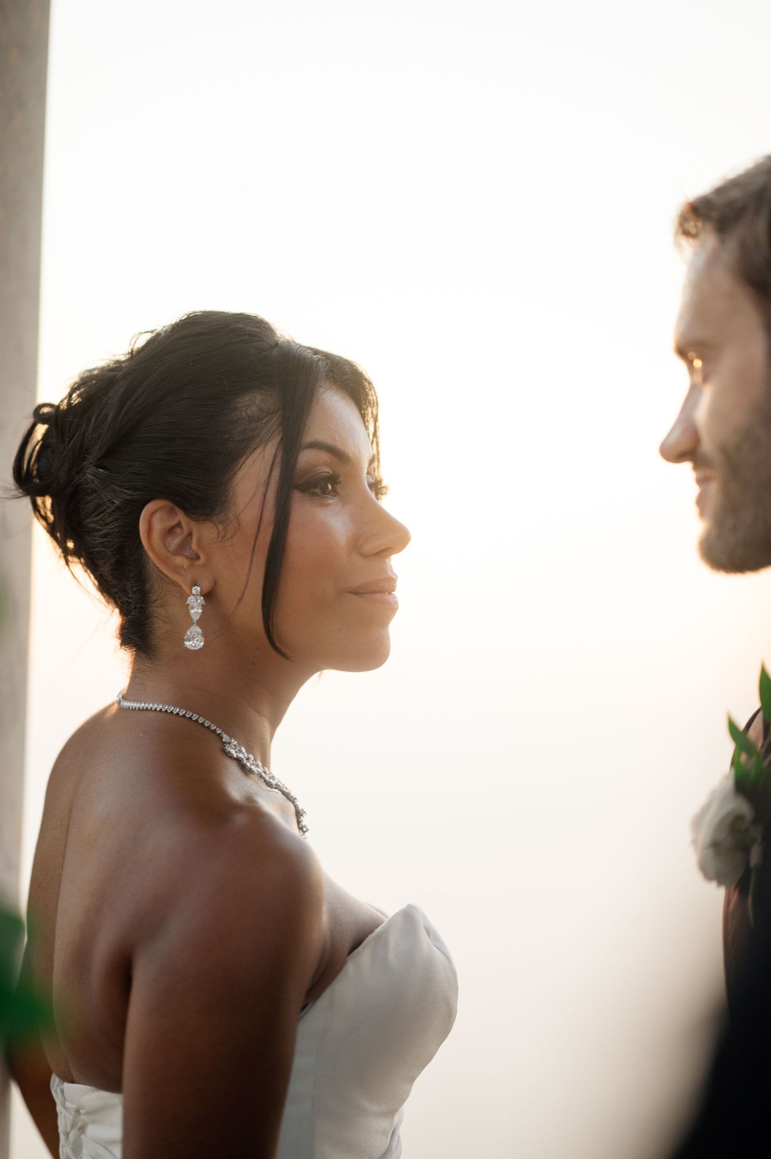 Beautiful bride with updo hairstyle and diamond jewelry gazes at groom in golden sunset light.