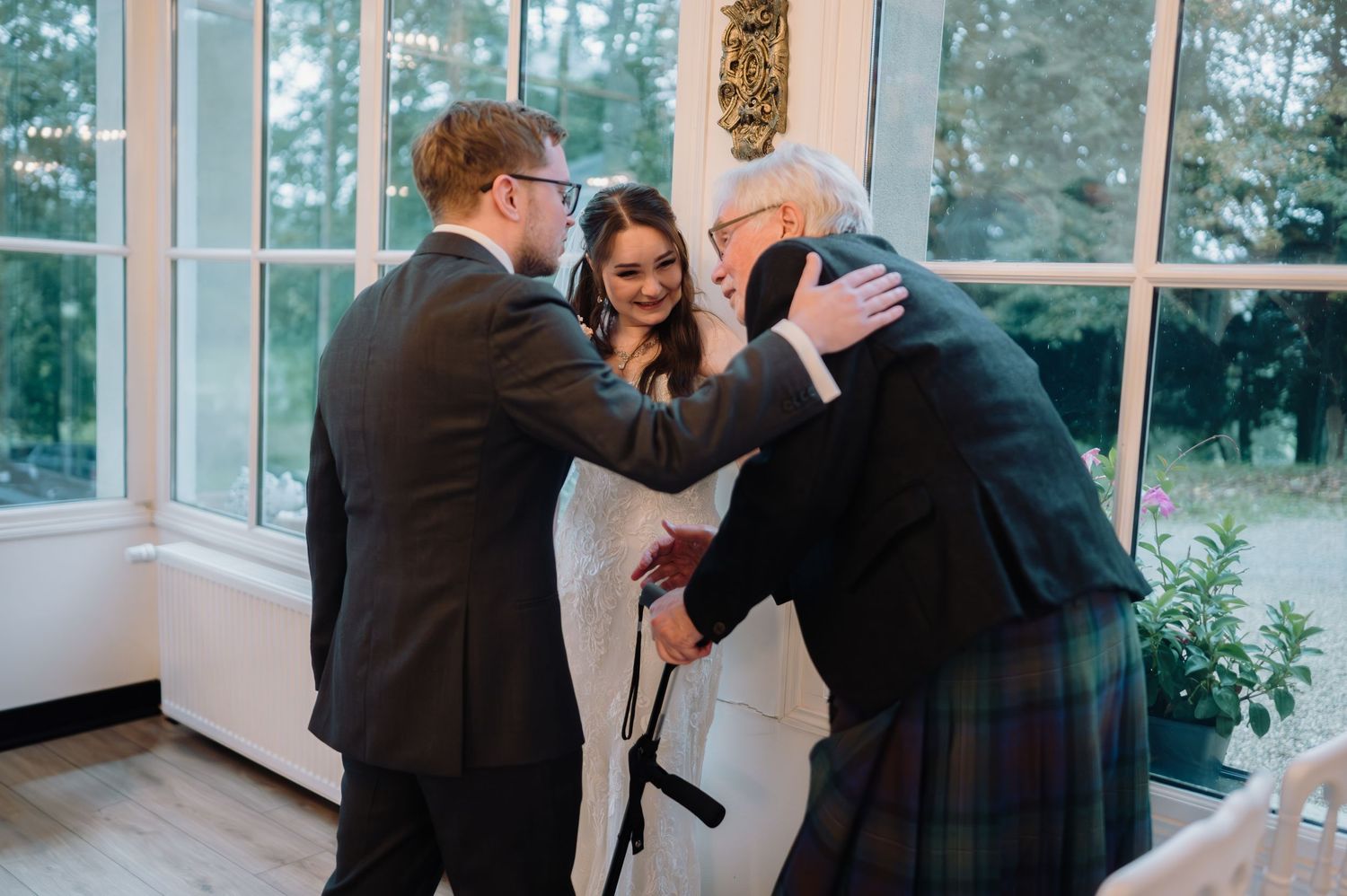 Groom and bride greeting elderly man in kilt at bright windowed venue during wedding celebration.