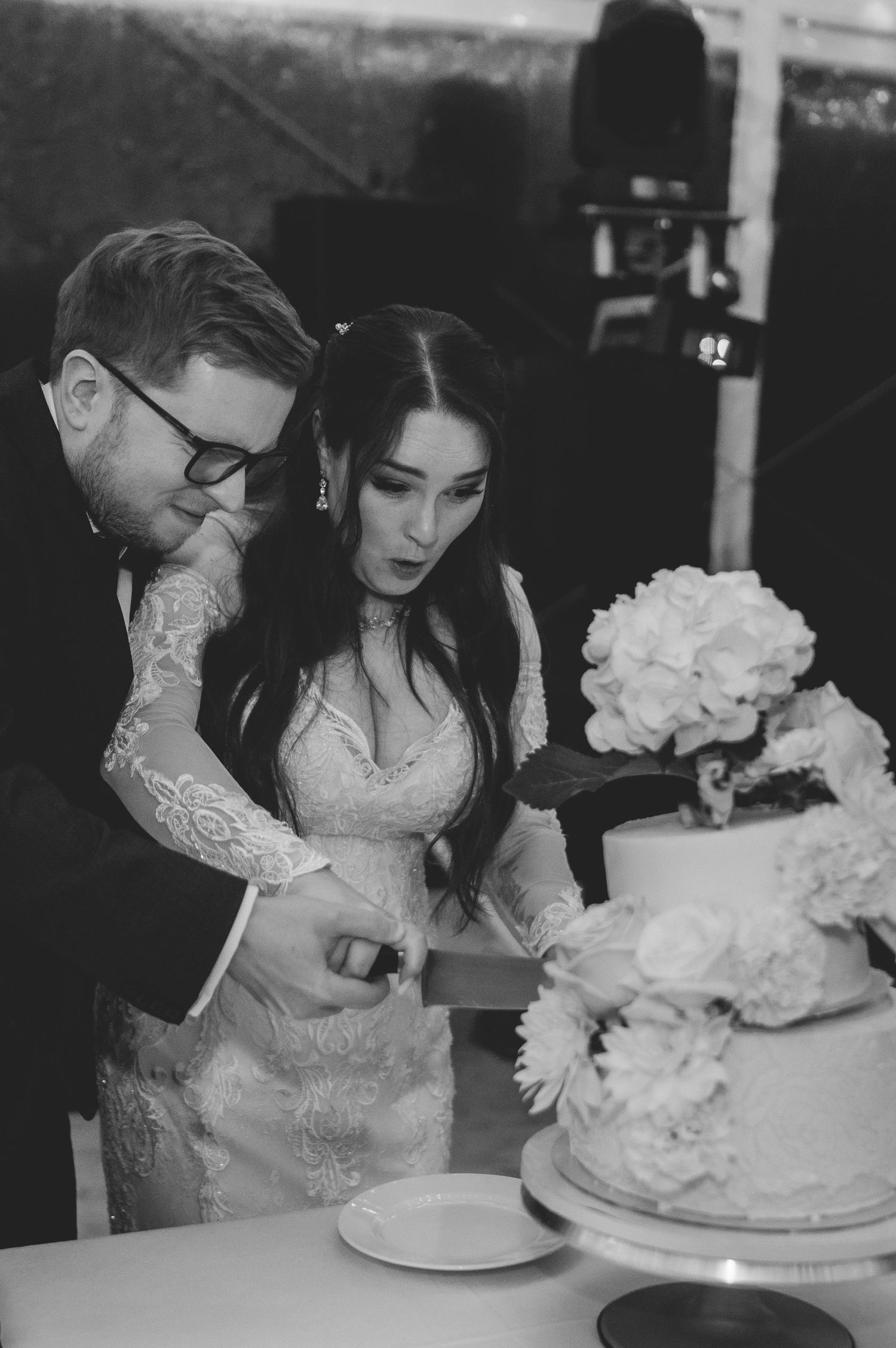 Black and white photo of bride and groom cutting their floral-decorated wedding cake together.