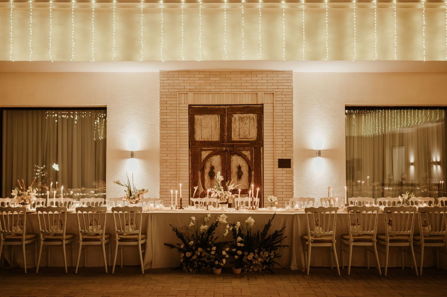 Warmly lit wedding reception hall with long banquet tables, candles, and ornate wooden doors.