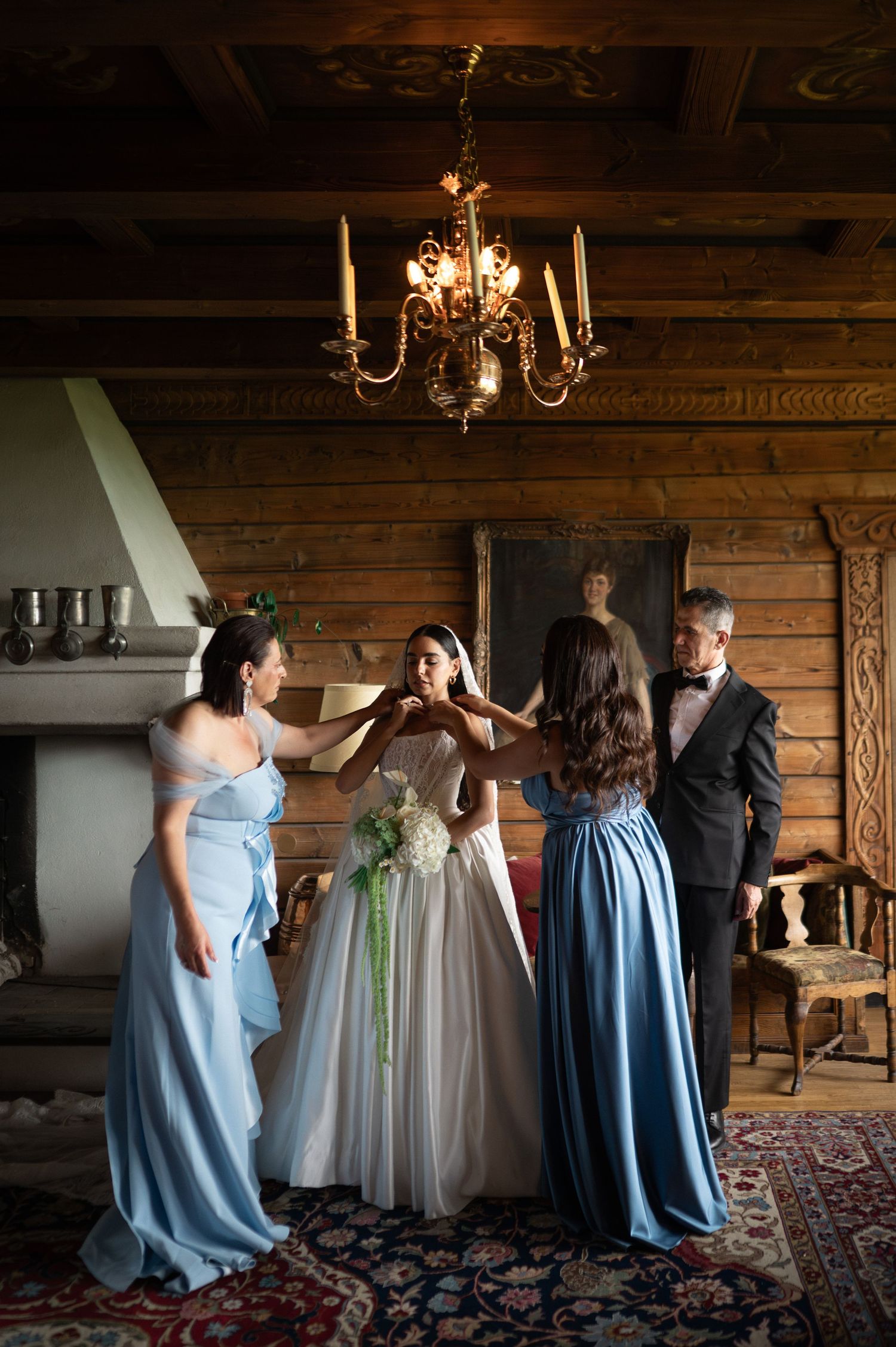 Bride surrounded by bridesmaids in blue dresses and groomsman in rustic wooden room with chandelier.