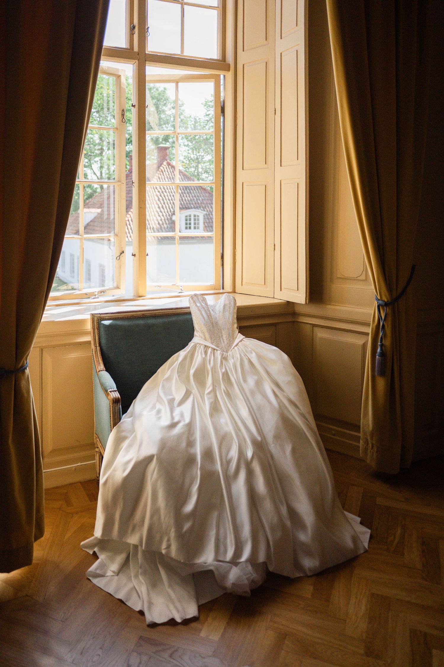 Elegant strapless ball gown wedding dress displayed on chair beside tall window with golden drapes.