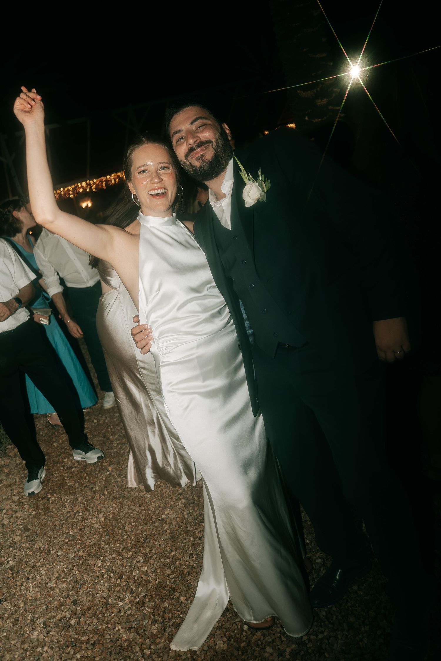 Joyful bride and groom dancing outdoors at night under string lights at wedding reception.