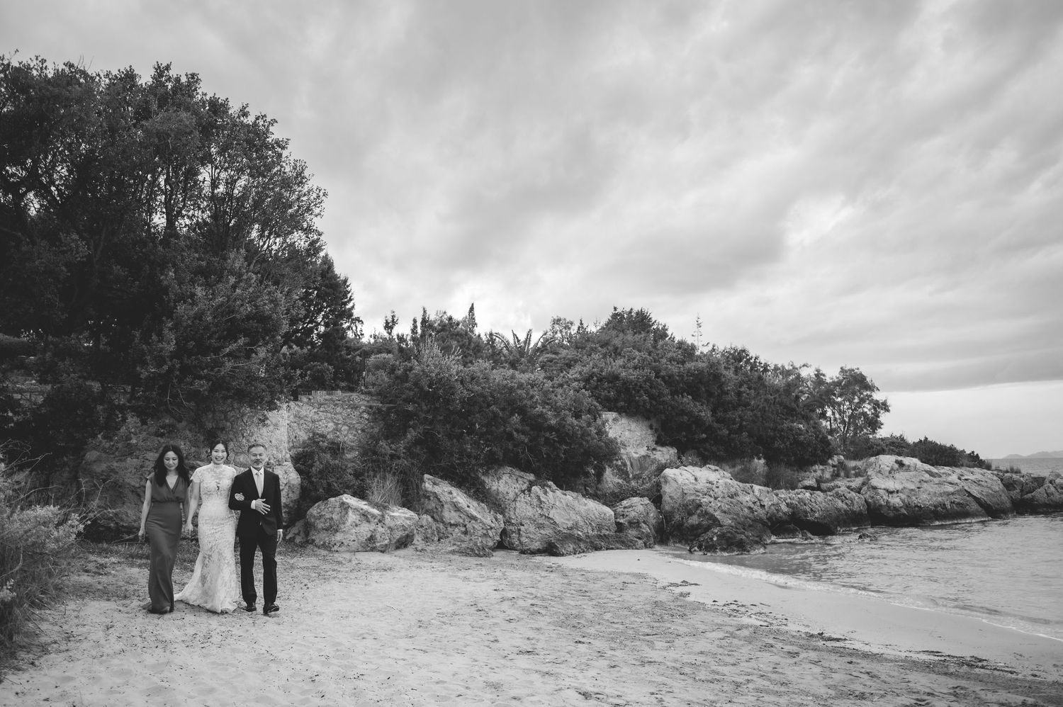 Black and white photo of wedding party walking along a sandy beach with rocky cliffs and trees.
