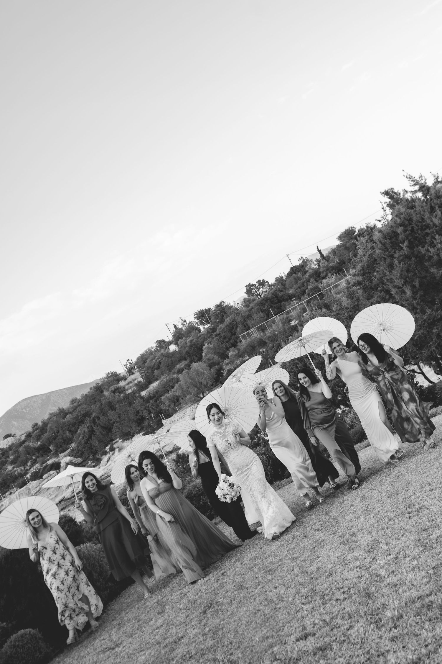 Bridal party posing with white parasols on a lawn with scenic hillside backdrop.