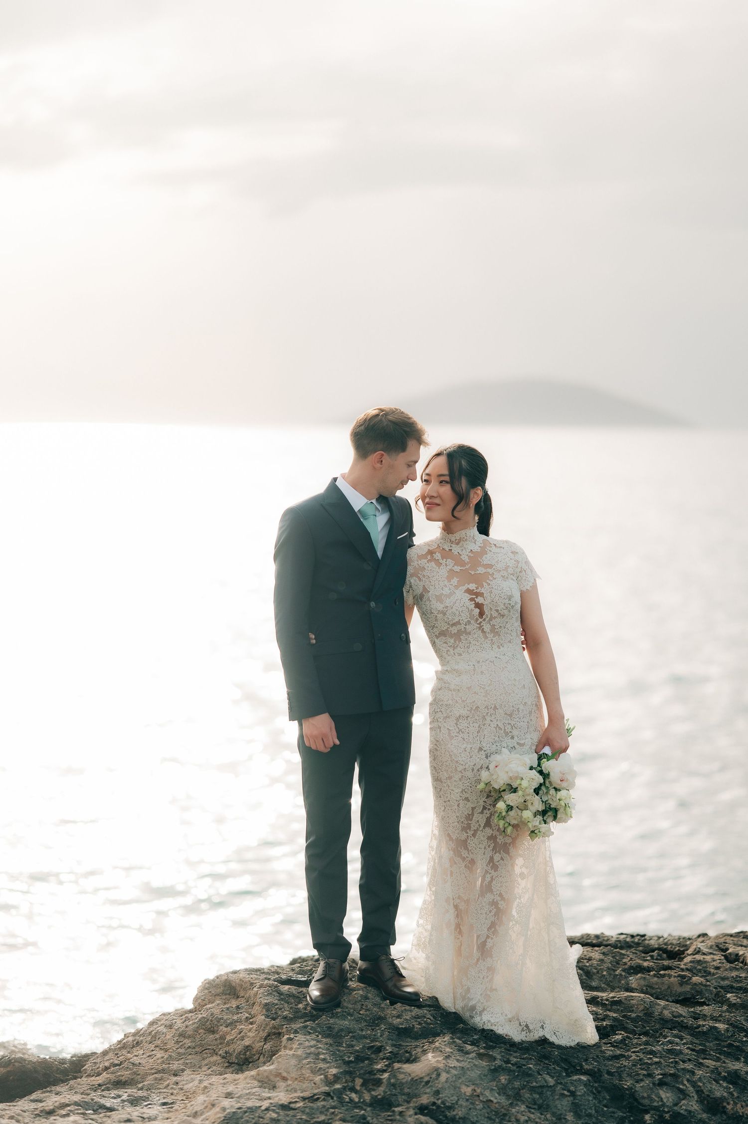 Elegant couple sharing a romantic glance on coastal rocks, bride in lace gown holding white bouquet at golden hour.