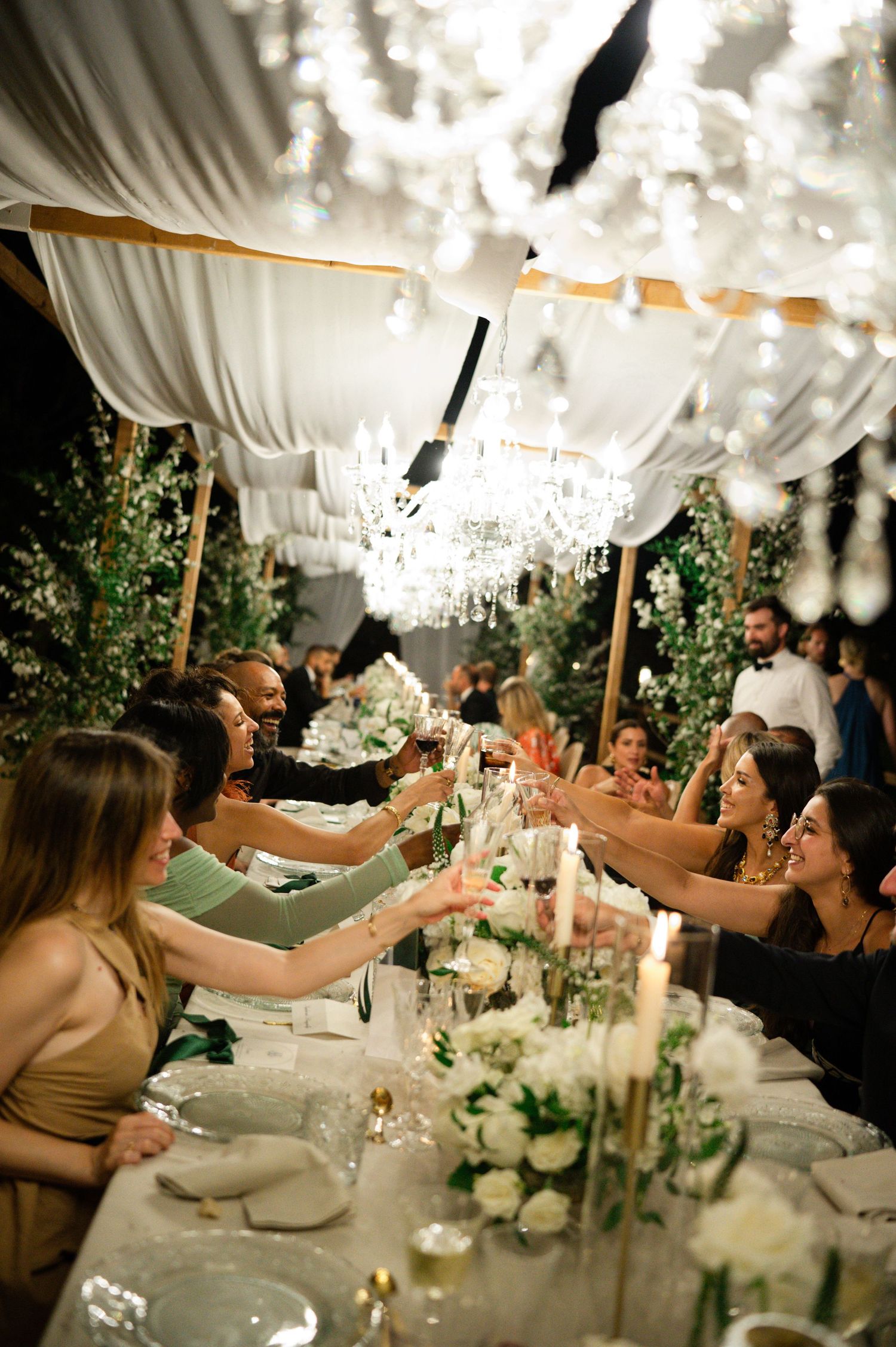 Elegant wedding reception guests toasting under crystal chandeliers with white floral centerpieces and candlelit long dining table.