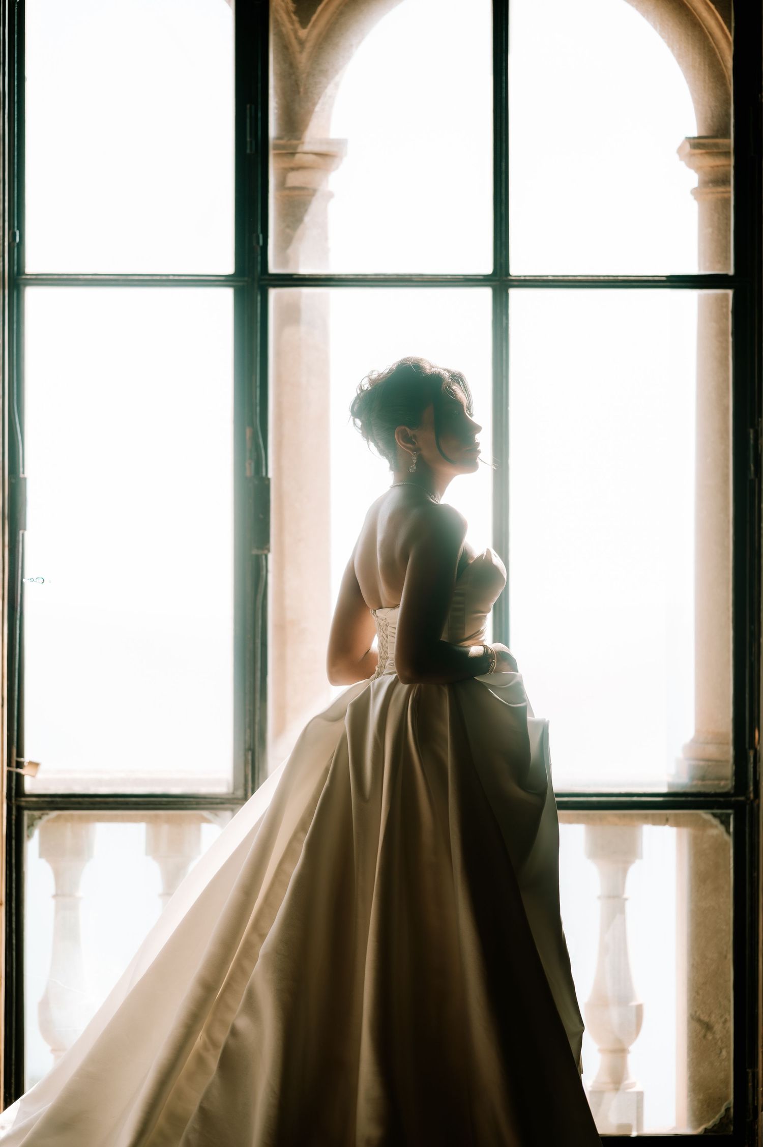 Backlit silhouette of bride in strapless white gown with flowing train standing at arched window.