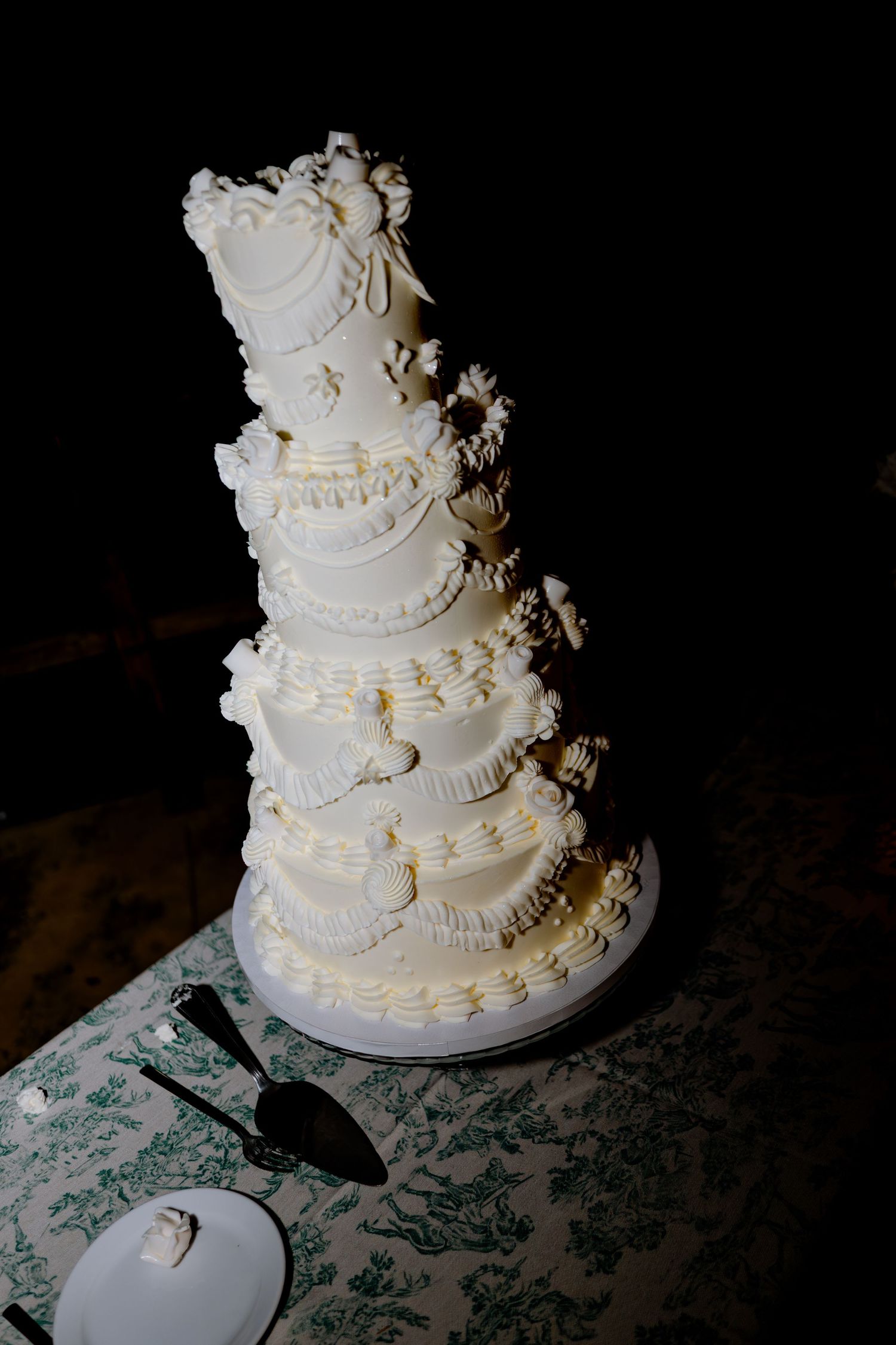 Elegant tall white multi-tiered wedding cake decorated with flowers and intricate piping on dark background.