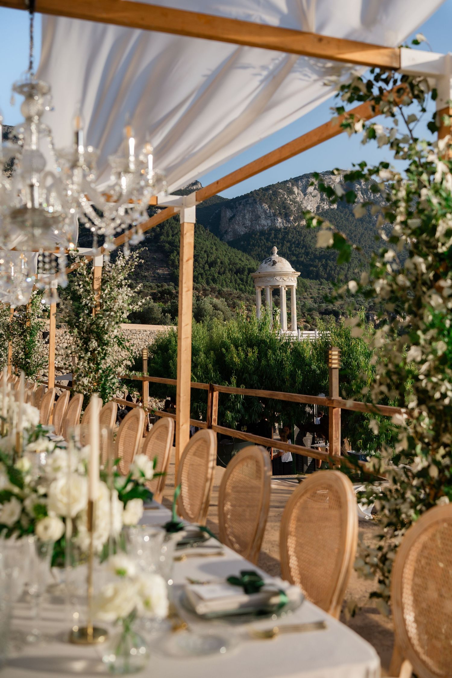 Wedding reception table with crystal chandelier, white floral arrangements, wicker chairs, and scenic mountain gazebo view.