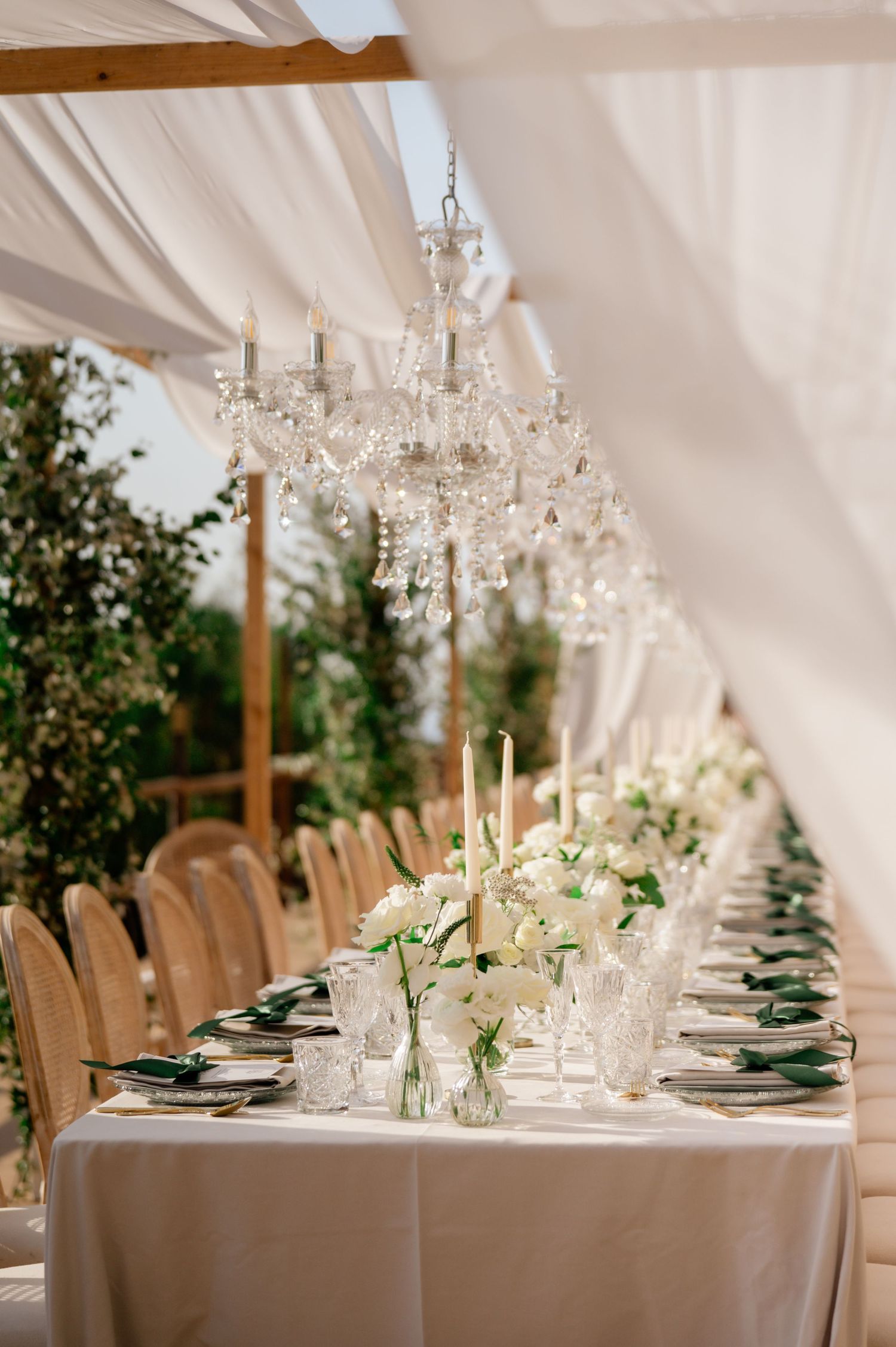 Luxurious wedding reception table with crystal chandeliers, white florals, candles and draped ceiling in elegant marquee.