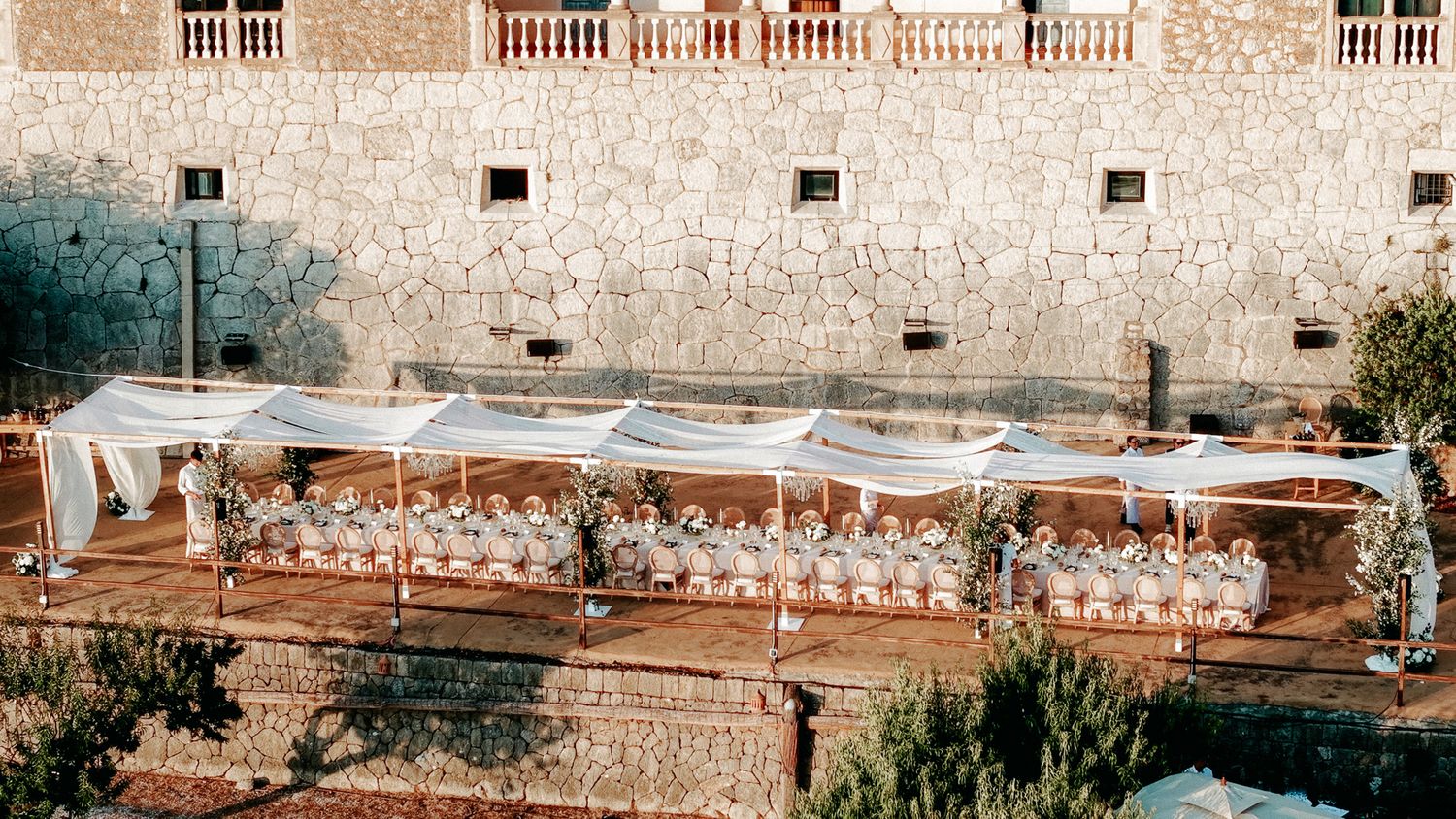 Bird's eye view of a sophisticated outdoor wedding banquet with white floral arrangements beside a historic stone wall.