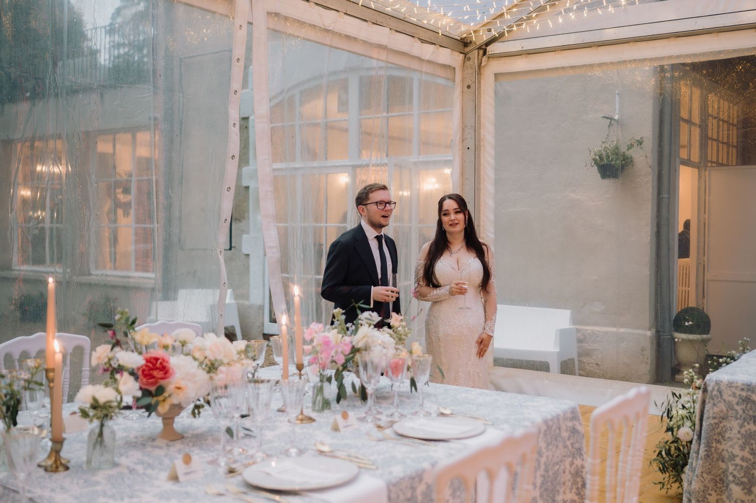 Bride and groom standing at a candlelit wedding reception table inside a glass greenhouse venue.