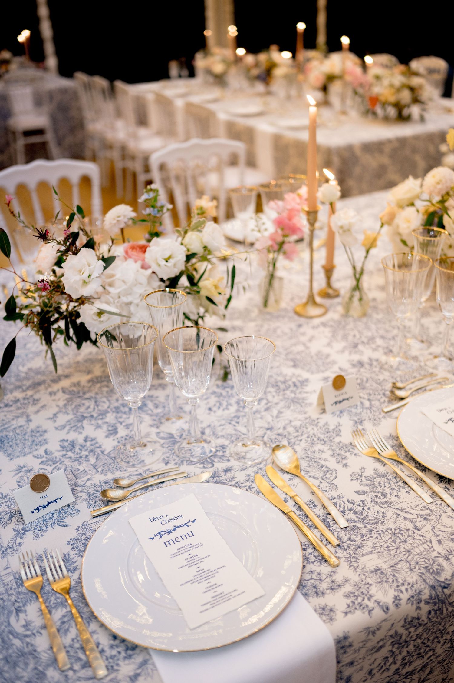 Wedding reception table with gold candlesticks, pink and white florals, gold flatware, and blue toile tablecloth.