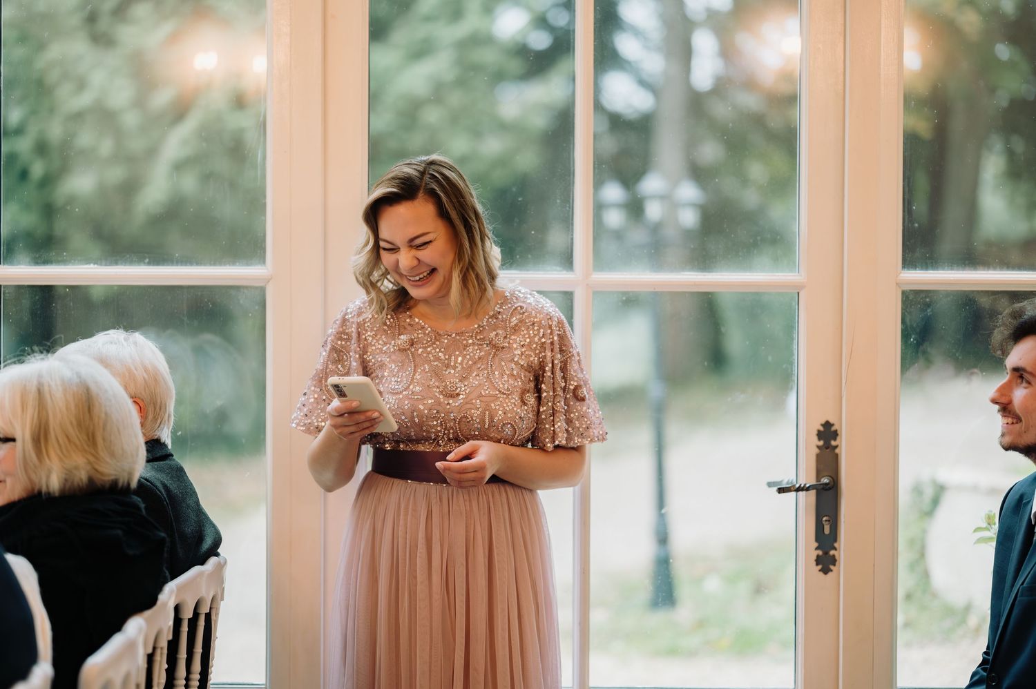 Smiling bridesmaid in blush sequin dress reads a heartfelt speech during wedding ceremony.