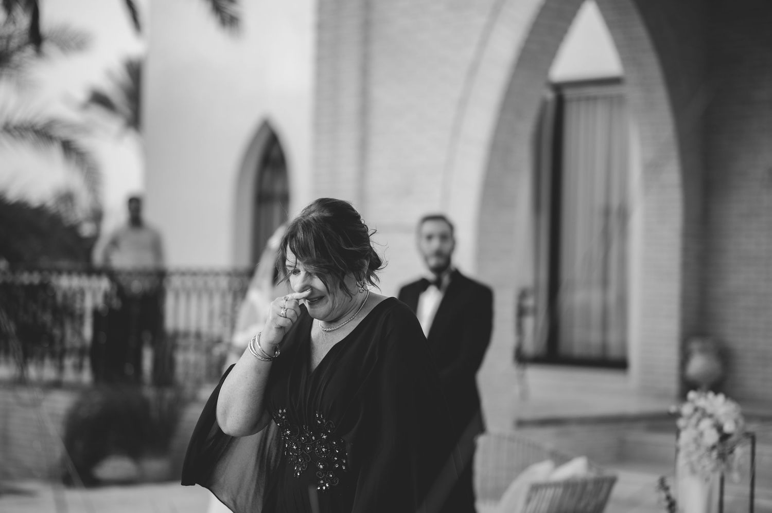 Emotional wedding guest wipes tears in black and white photo outside elegant arched ceremony venue.